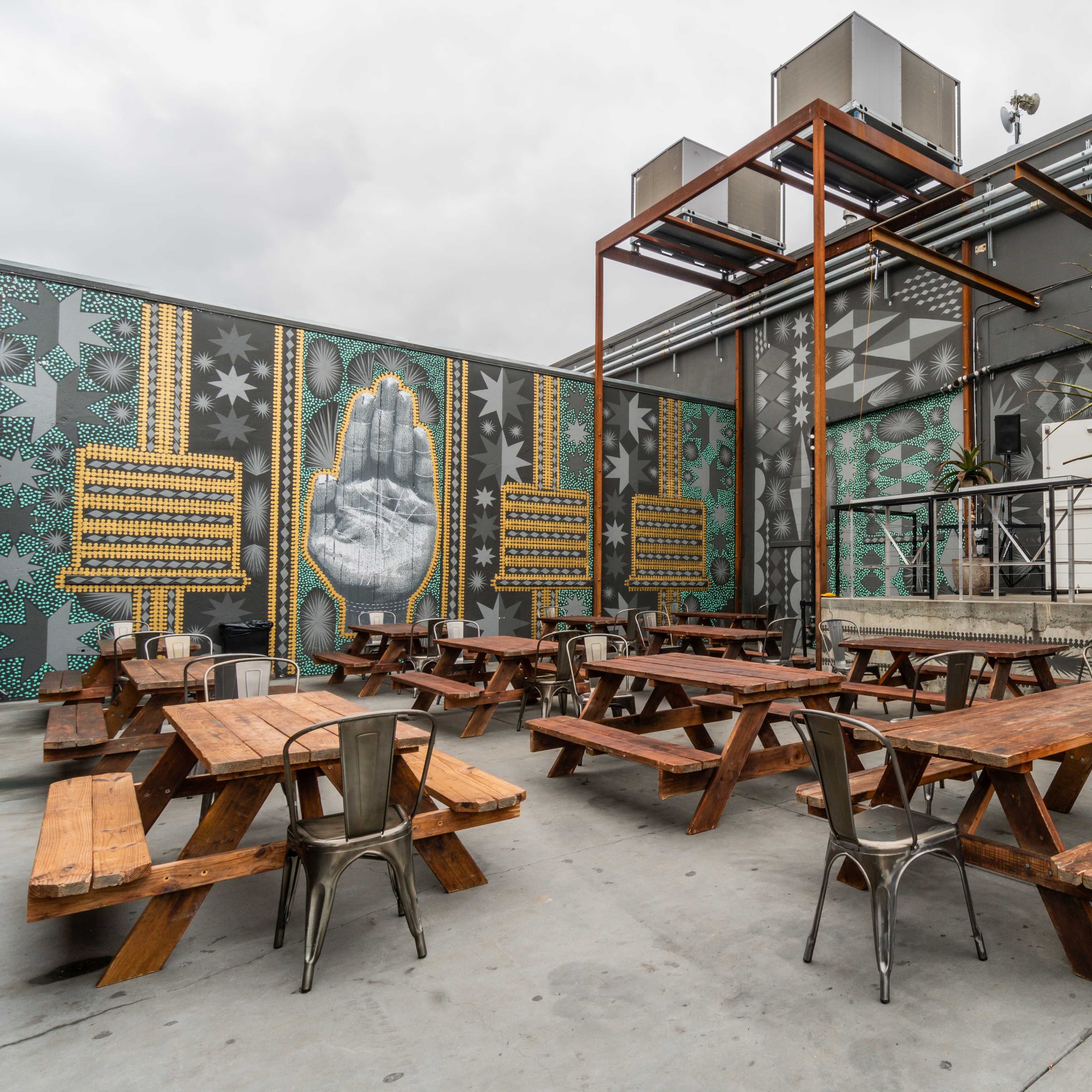 An outdoor seating area with multiple wooden picnic tables and a palm tree, set against a colorful mural on a concrete wall.
