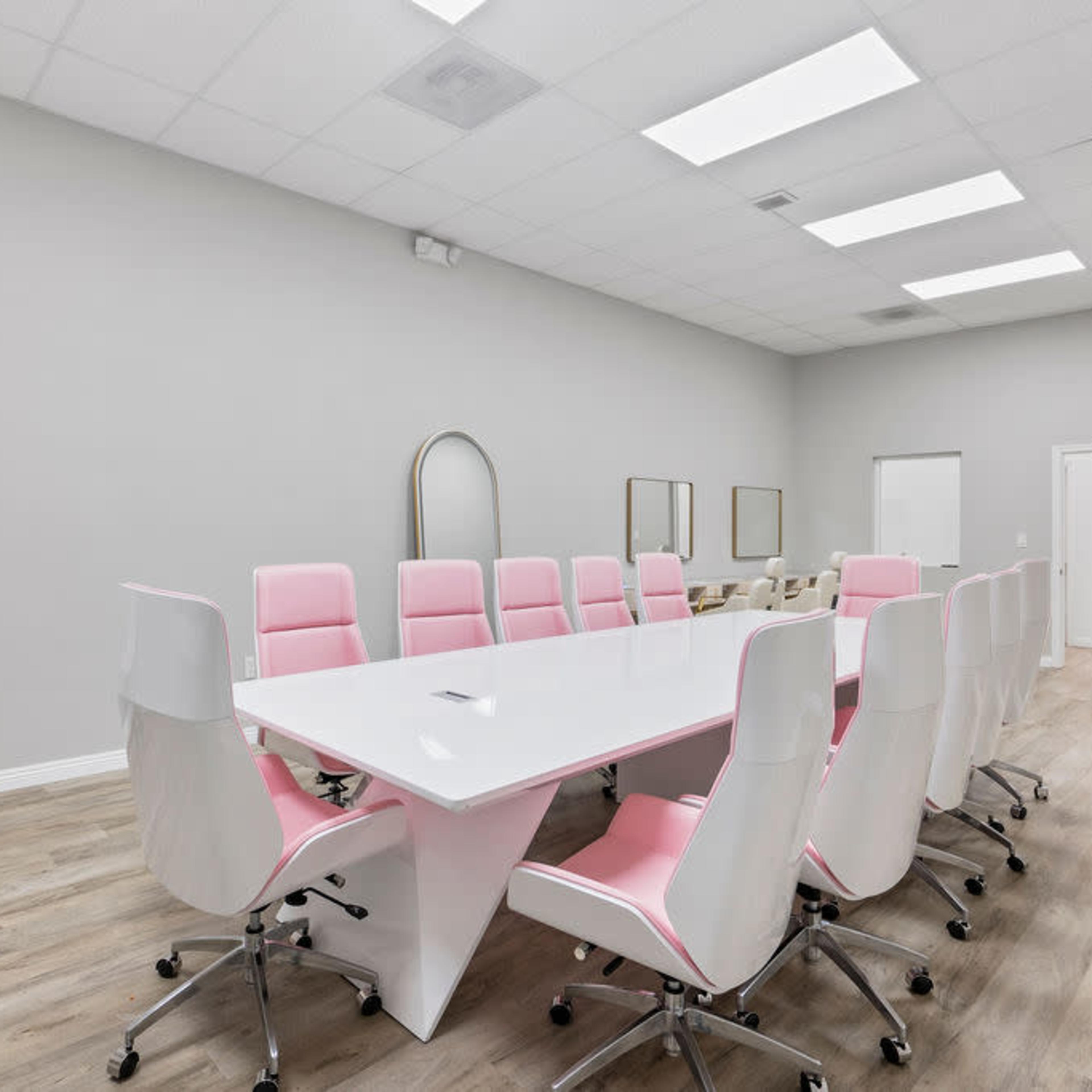 A modern conference room features a large white table surrounded by pink and white chairs, with large windows and minimalist decor.