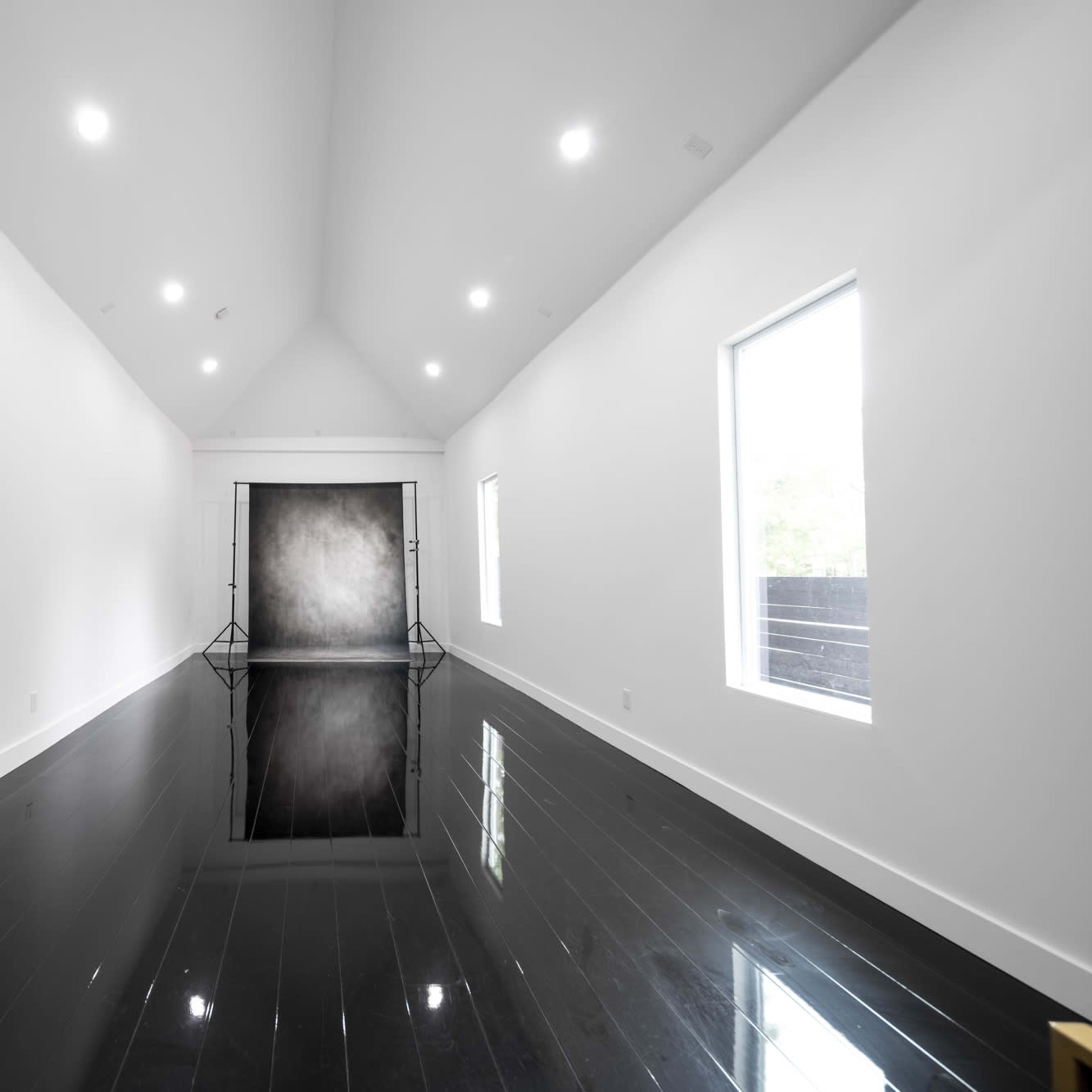 A spacious, brightly lit room with white walls, large windows, and a glossy black floor reflecting a backdrop setup at the far end.
