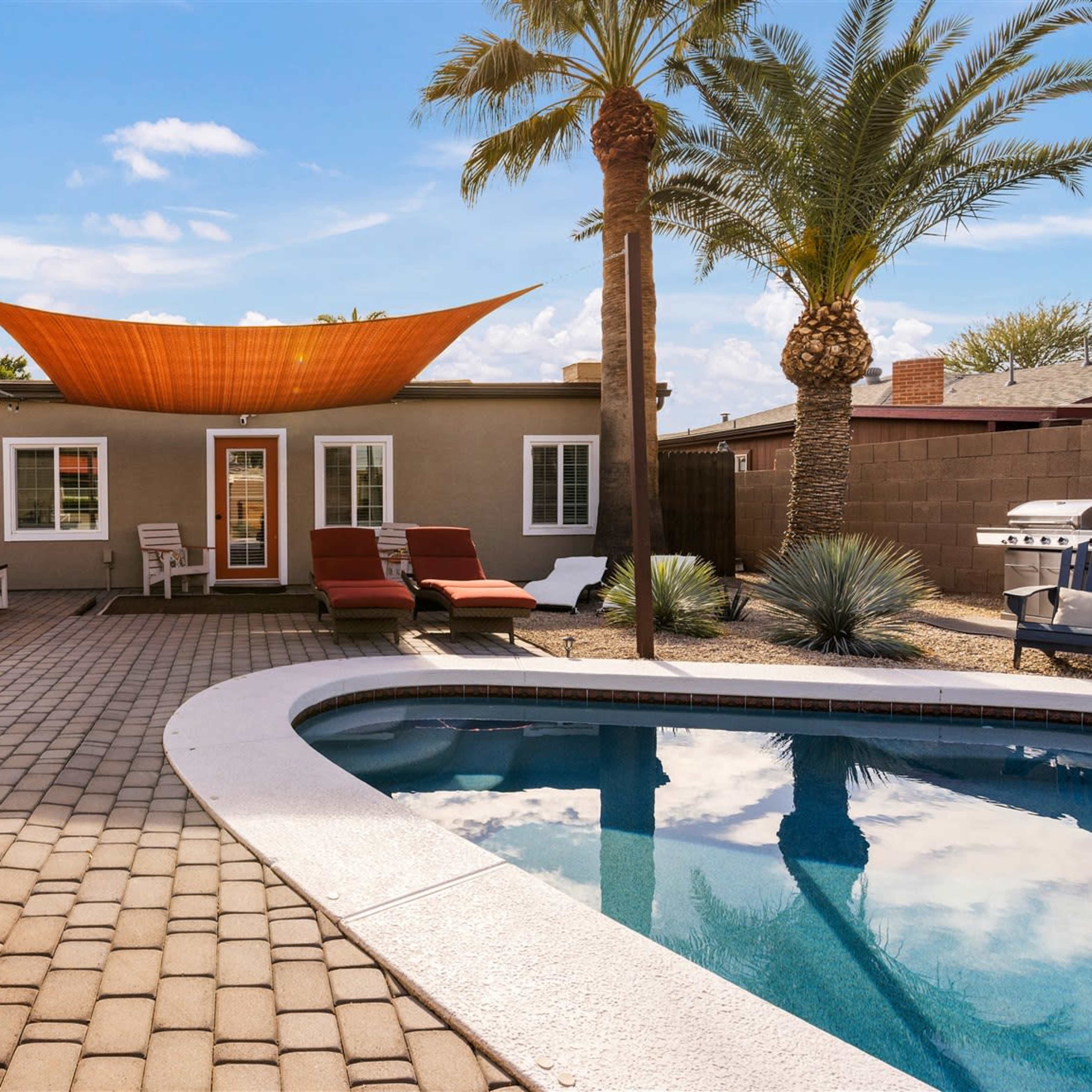 A backyard featuring a swimming pool, patio area with lounge chairs, artificial grass, and a shaded seating area, bordered by palm trees and a fence.