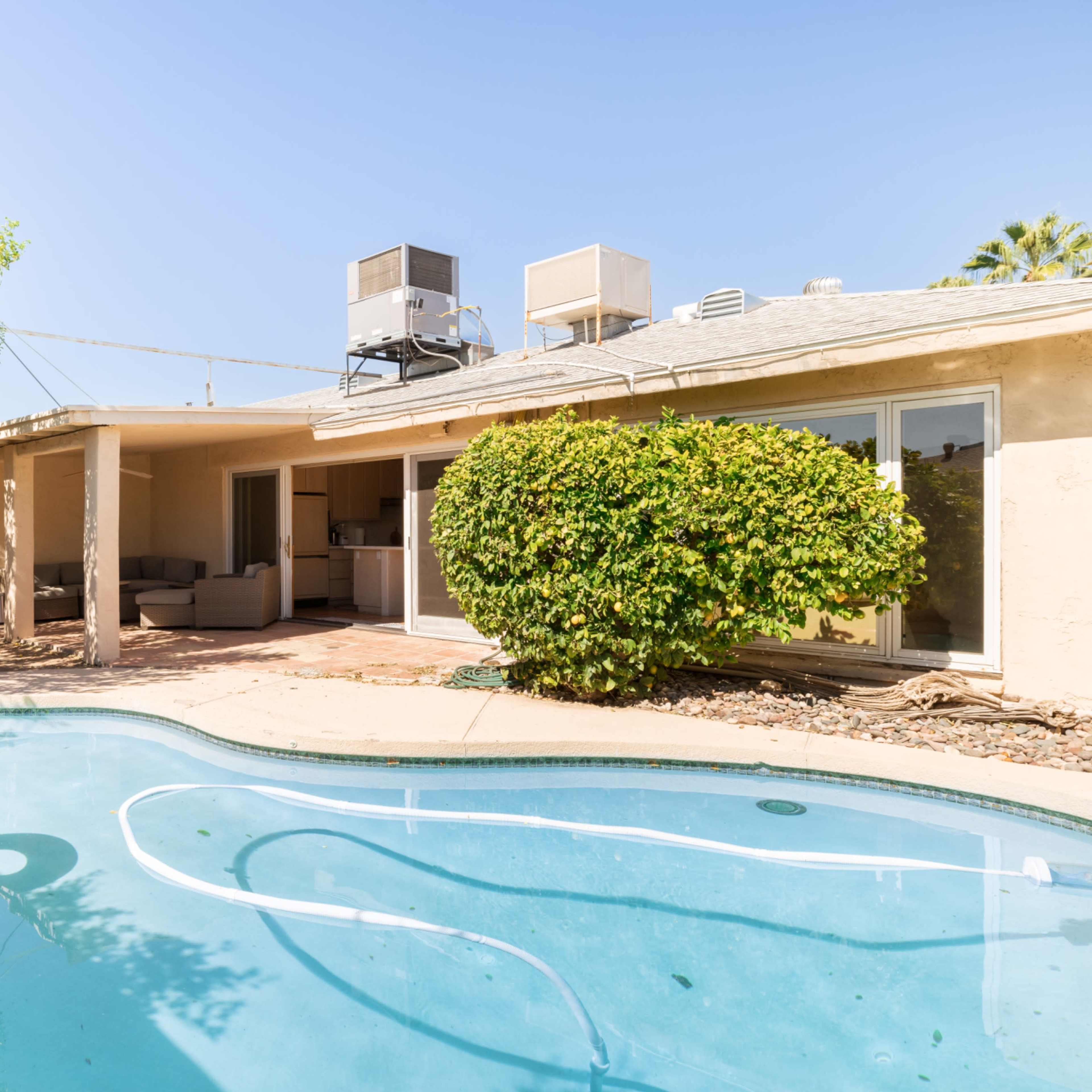 A backyard features a swimming pool with a pink float, surrounded by greenery and a single-story house with exposed air conditioning units on the roof.