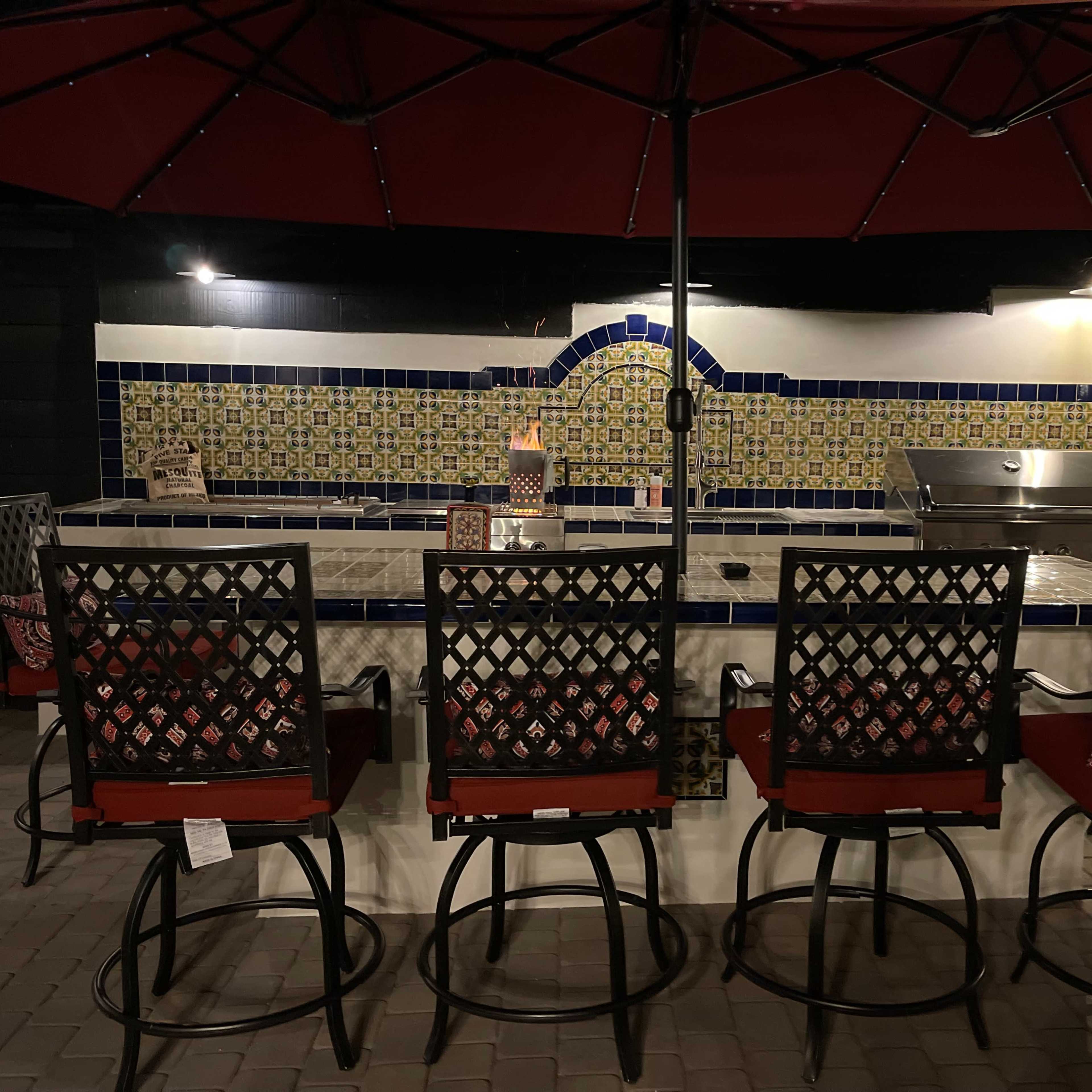 An outdoor kitchen area with a tiled backsplash, a grill, and six bar stools arranged around a counter, all under a red umbrella.