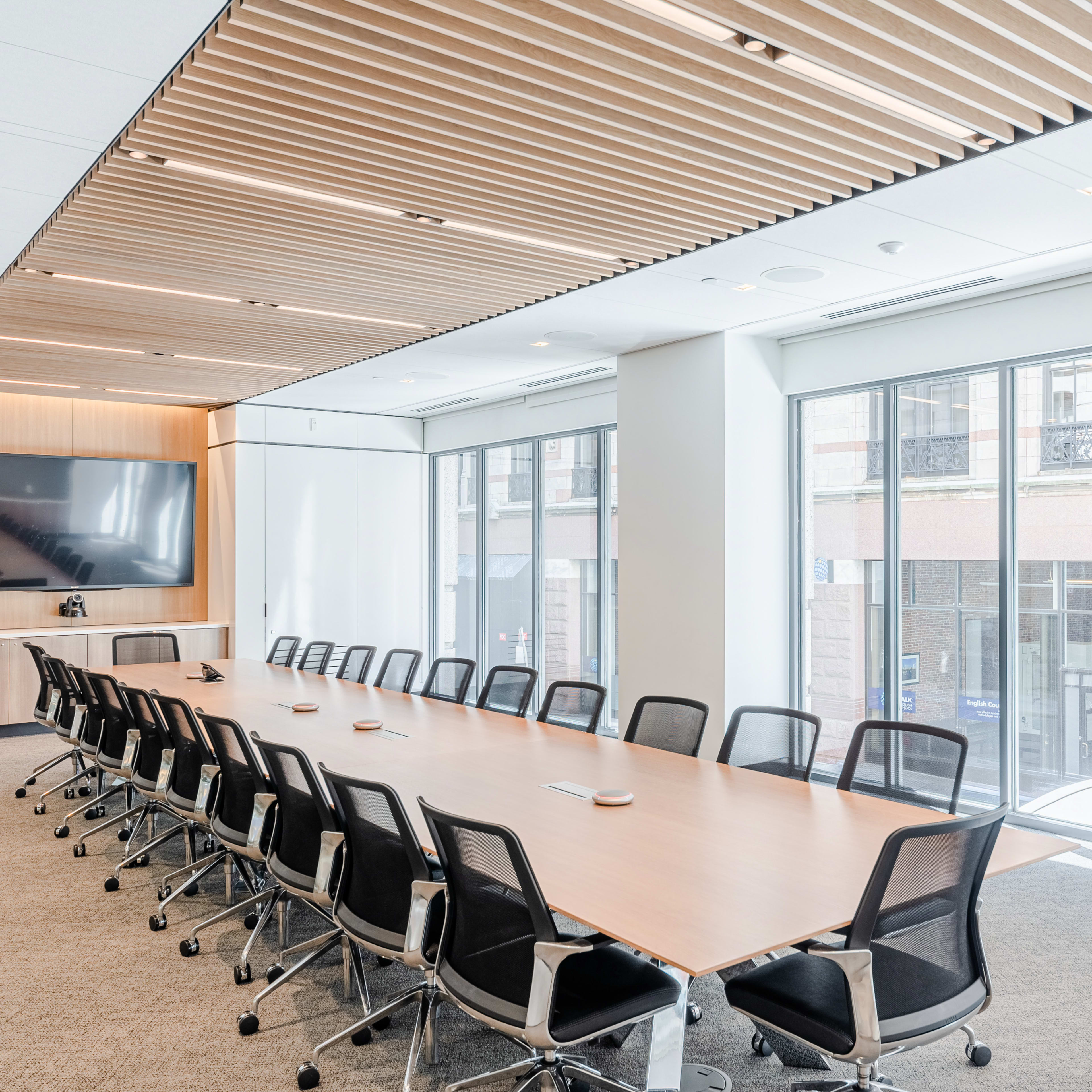 A long conference table with black chairs is set up in a modern meeting room featuring large windows and a wall-mounted screen.