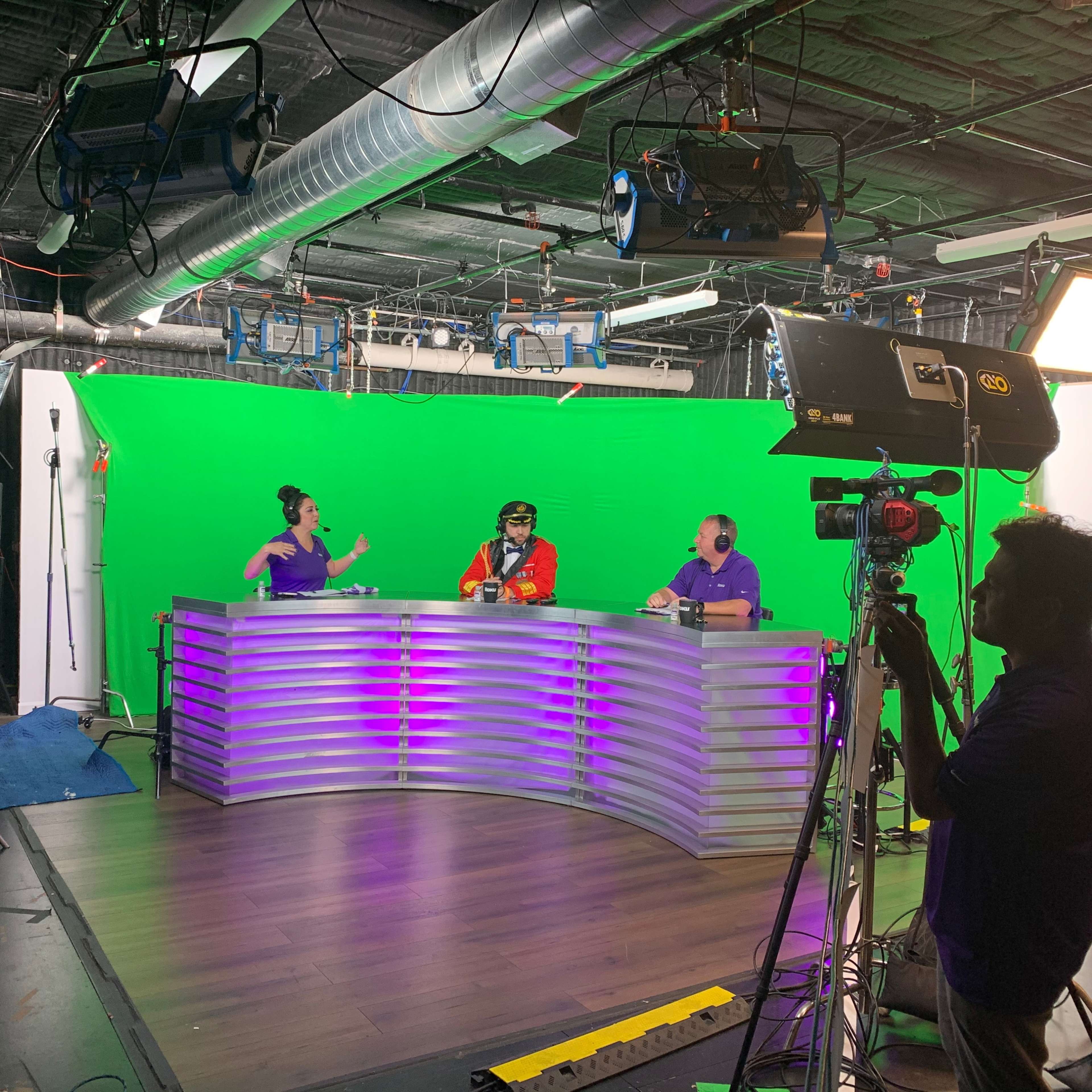 Two hosts sit at a curved desk in a studio with a green screen backdrop, while a cameraman operates a camera nearby.