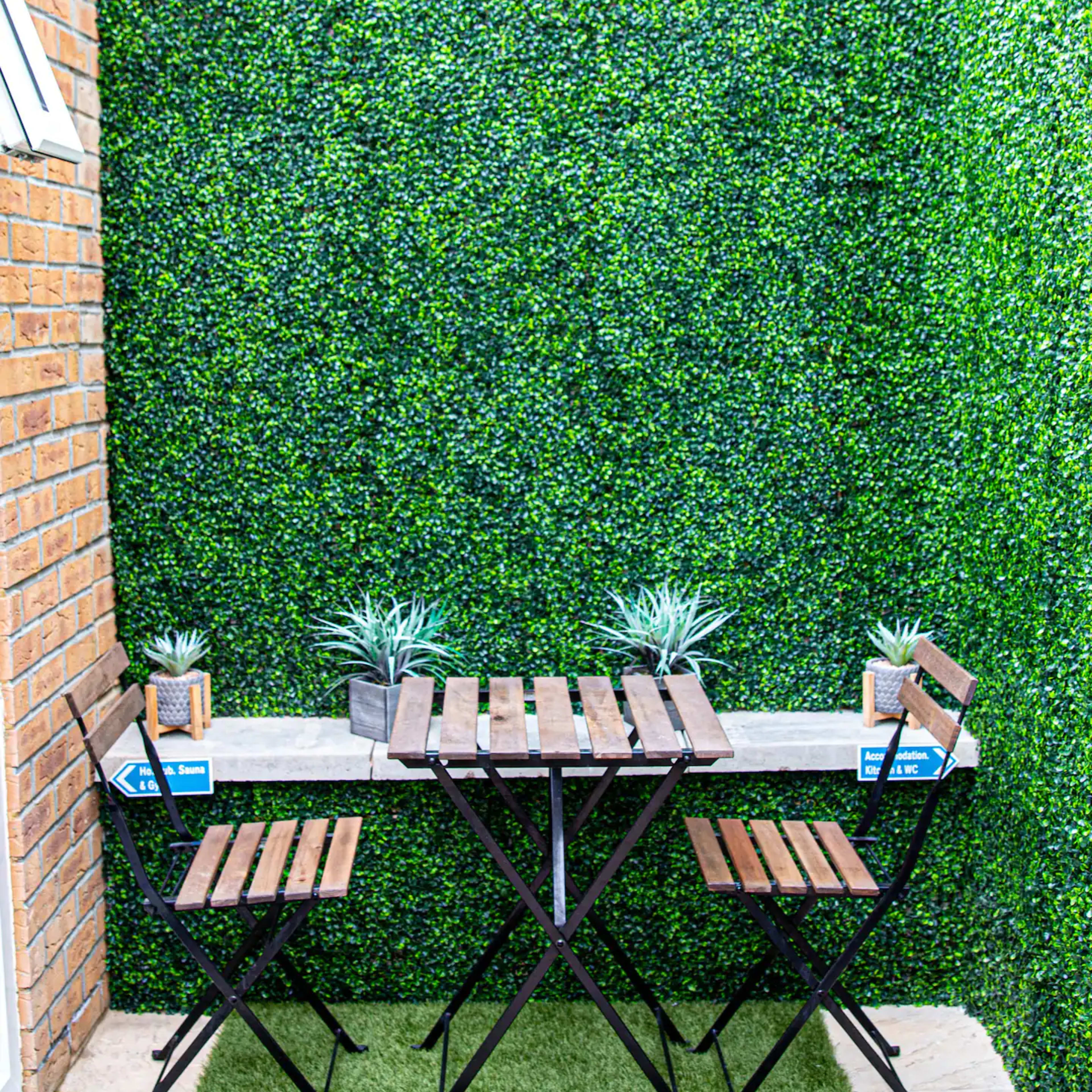 A small outdoor seating area features a wooden table with two folding chairs and potted plants against a green wall of artificial foliage.