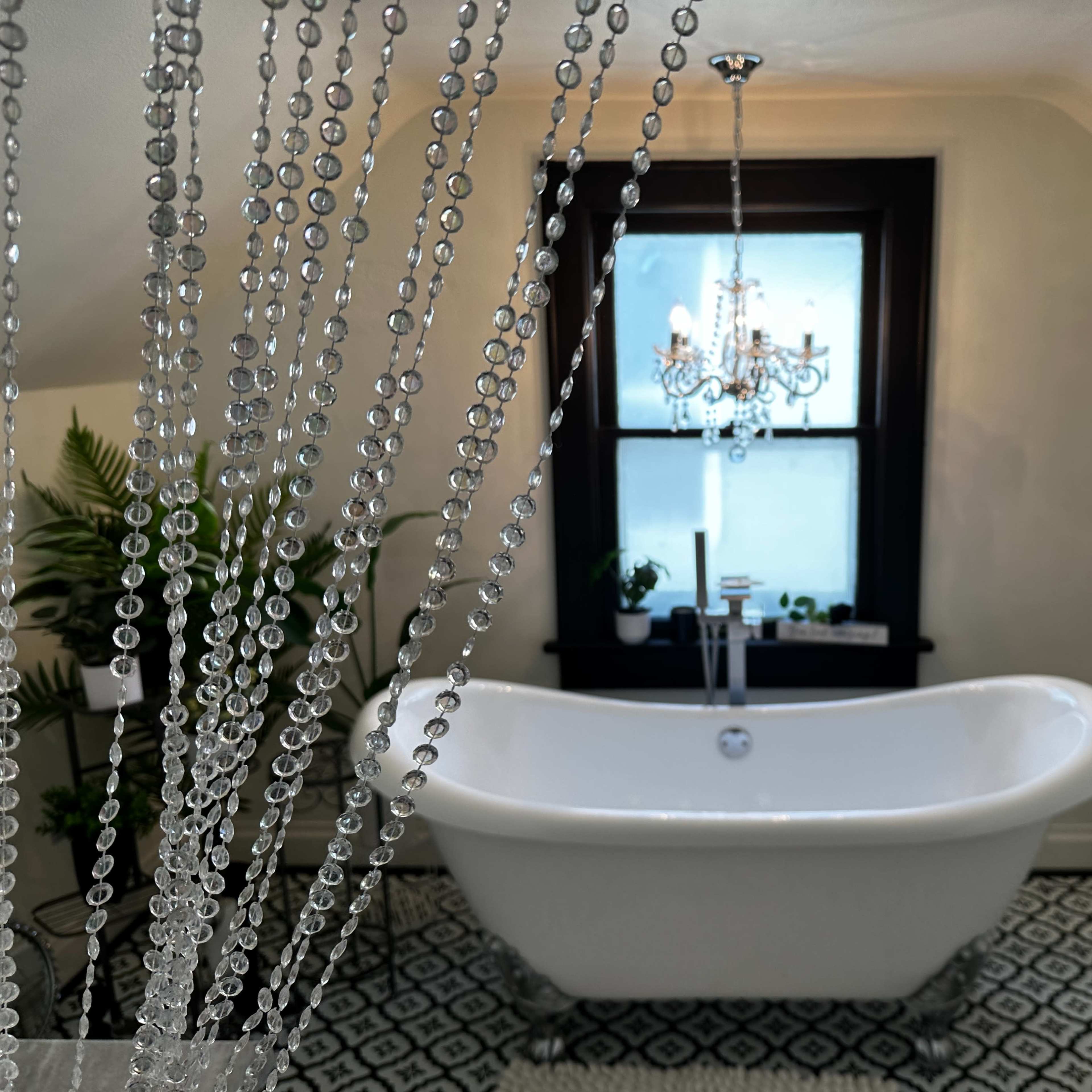A freestanding white bathtub is positioned in a bathroom adorned with a chandelier, surrounded by string curtains made of beads, with a window revealing a view of indoor plants.