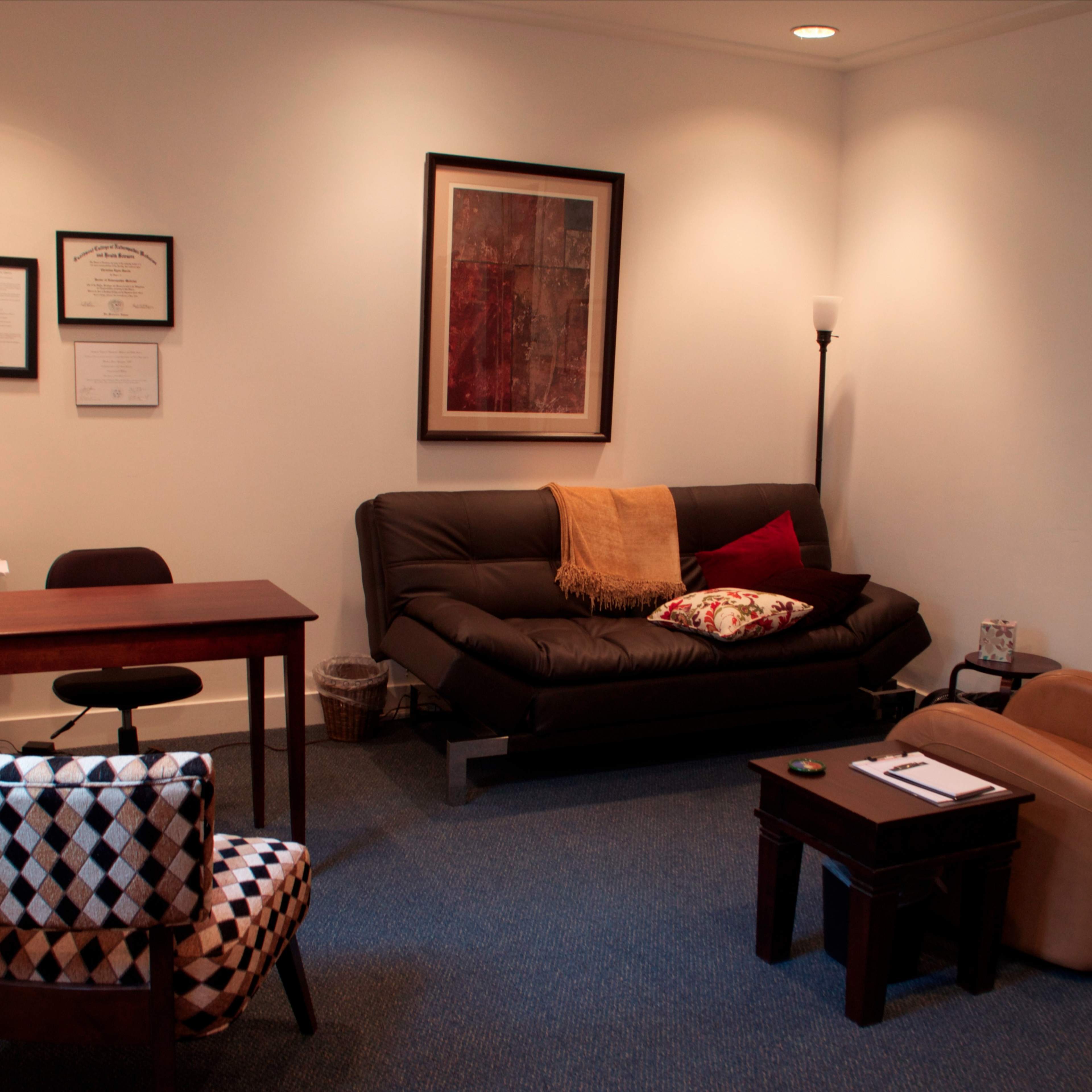 A simple office space featuring a black sofa, a desk with a chair, and a brown armchair, all set against white walls with framed artwork and certificates displayed.