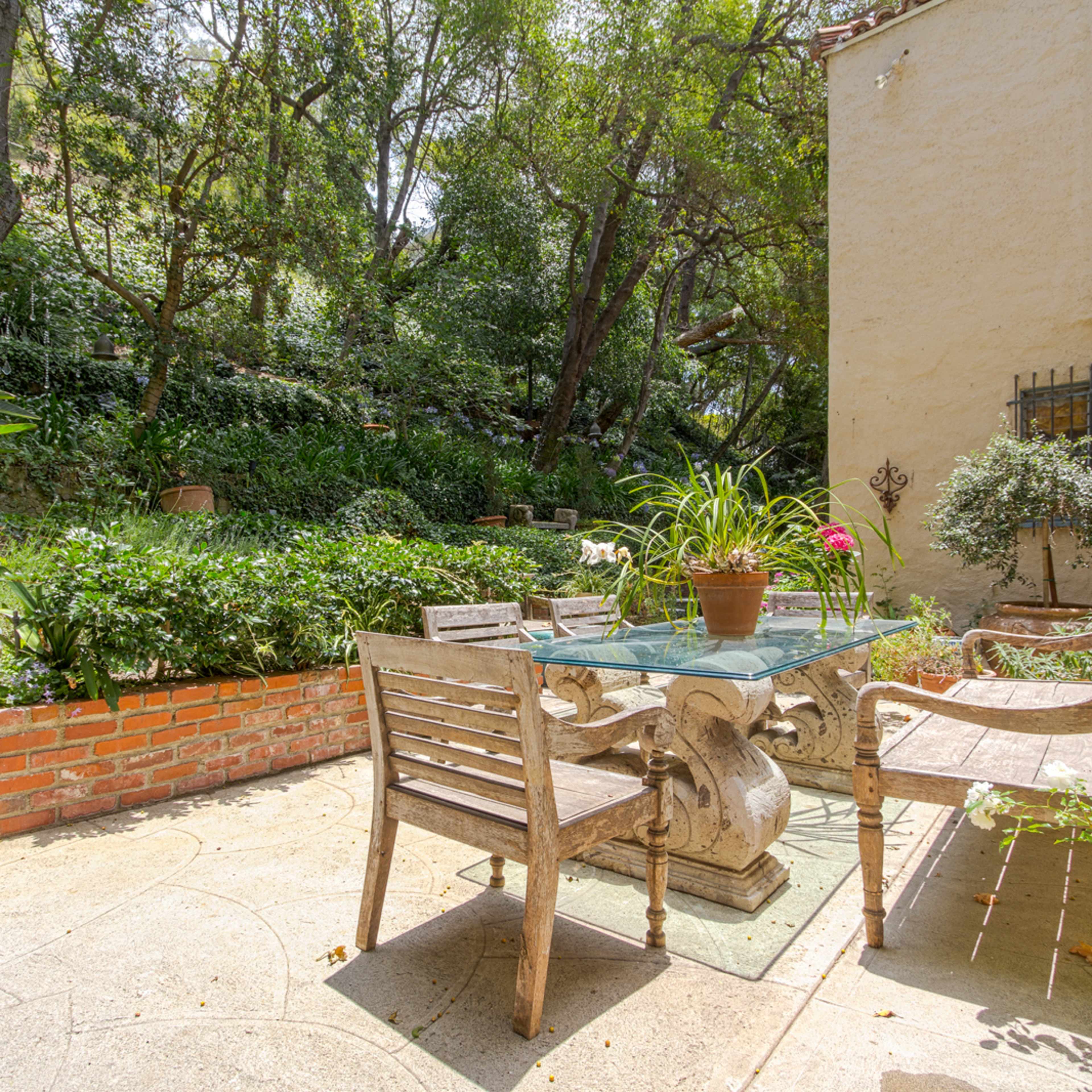A stone patio features a glass-top table surrounded by wooden chairs, nestled among lush greenery and steps leading to a higher garden area.
