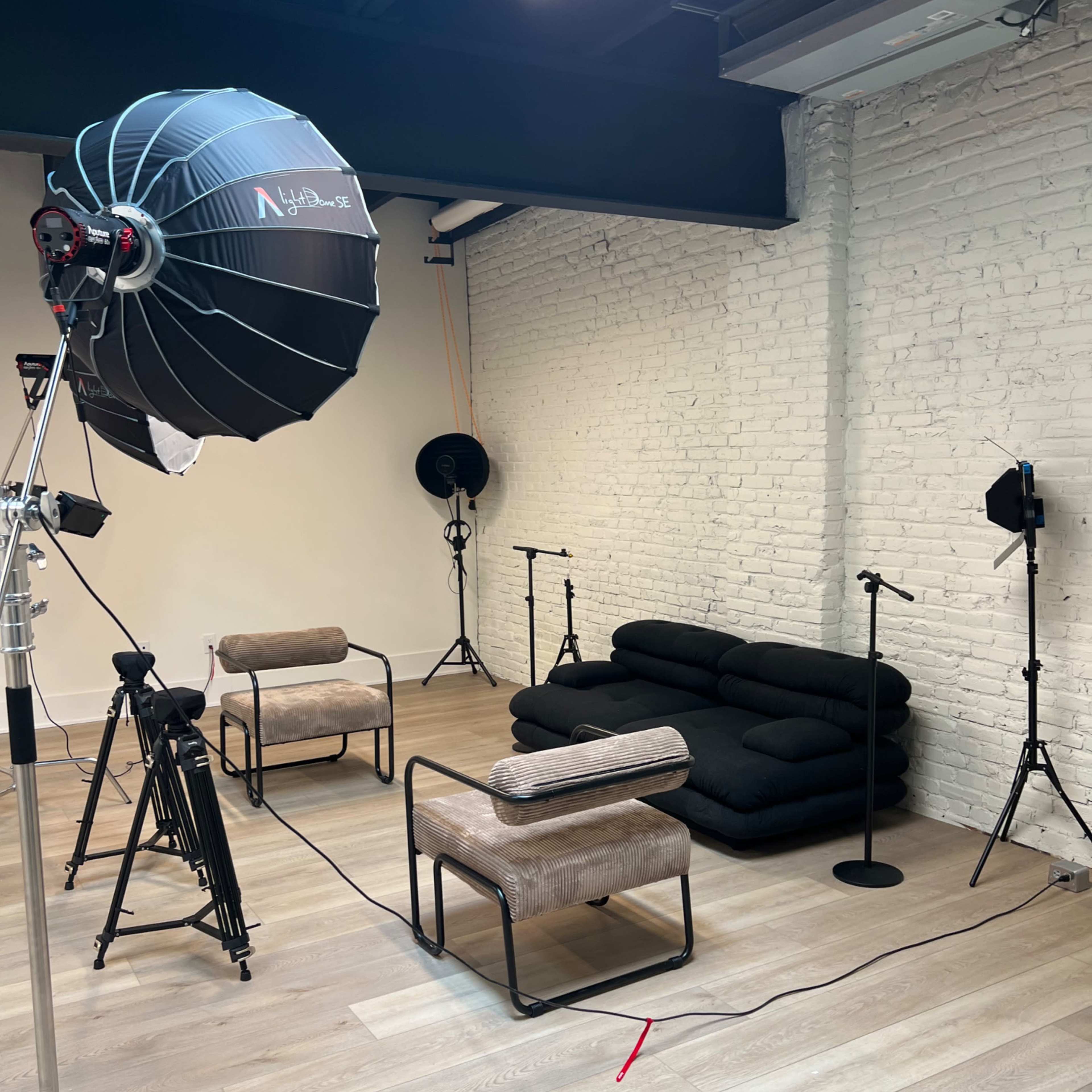 A minimalist photo studio setup with a sofa, two chairs, and several lighting and microphone stands on a wooden floor.