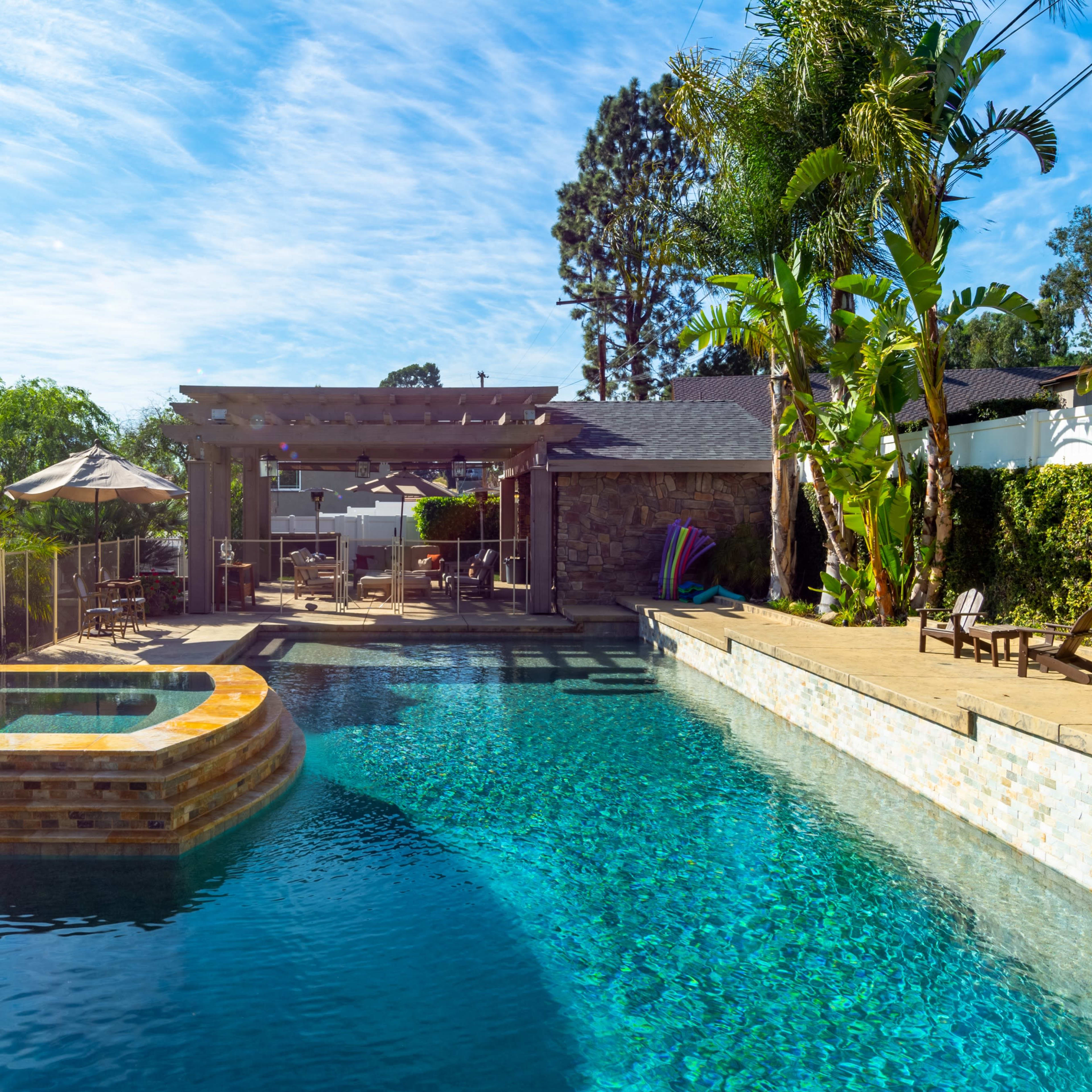 A rectangular swimming pool with a connected spa, surrounded by landscaped greenery, outdoor seating, and a pergola.