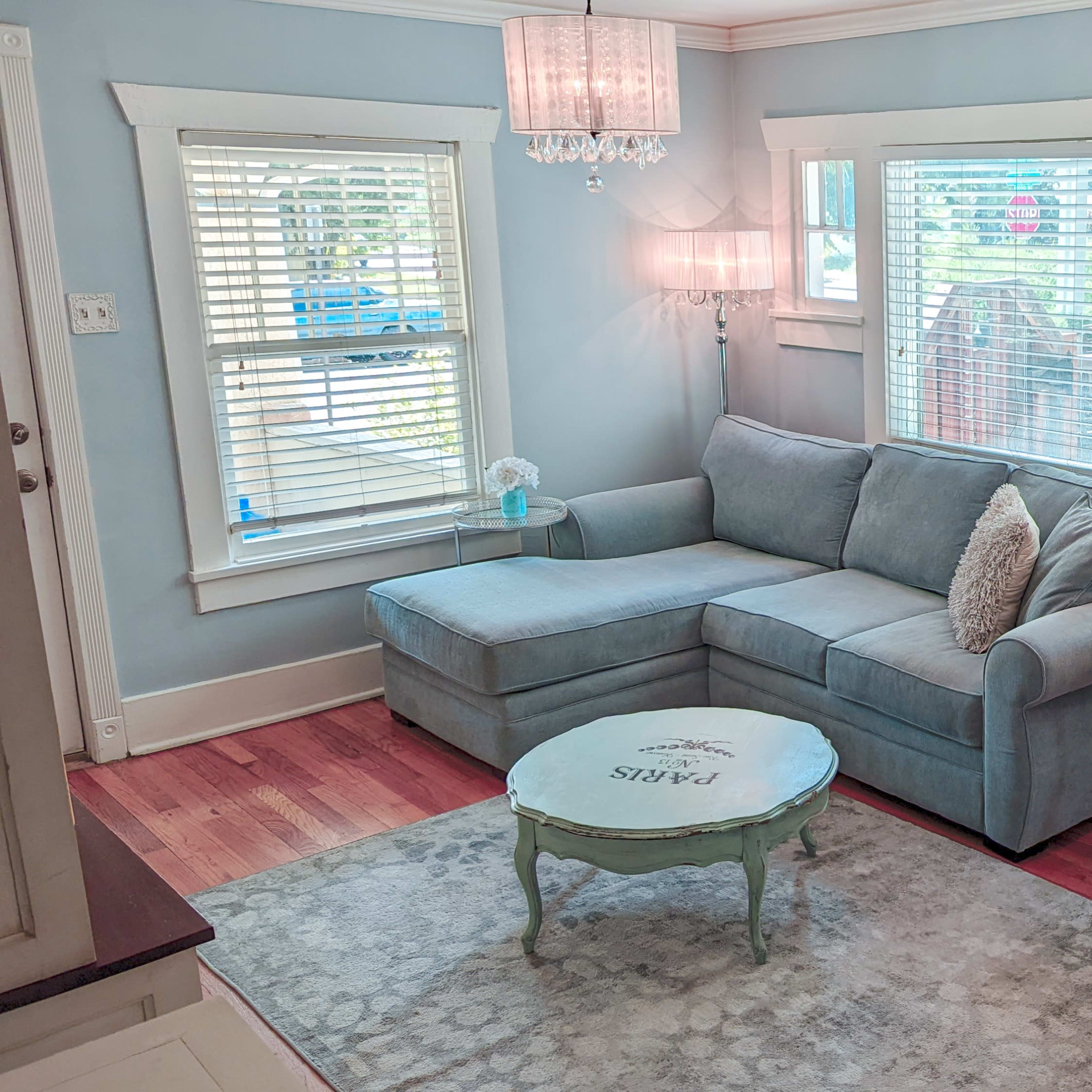 A light blue living room featuring a sectional sofa, a small round coffee table, a lamp, and a decorative armchair, all illuminated by natural light.