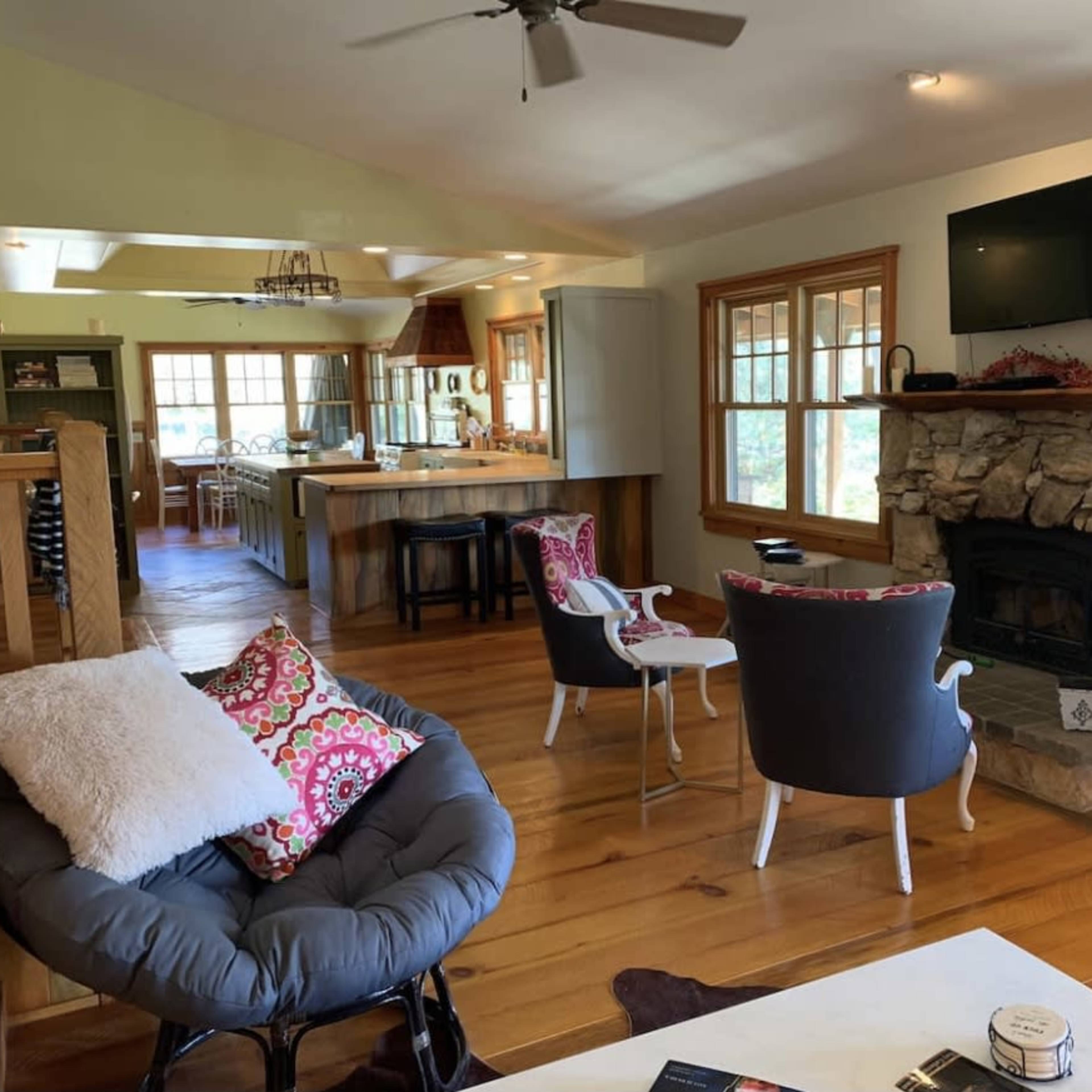 A spacious living area with wooden flooring, a stone fireplace, and comfortable seating arranged around a coffee table, leading to a kitchen in the background.