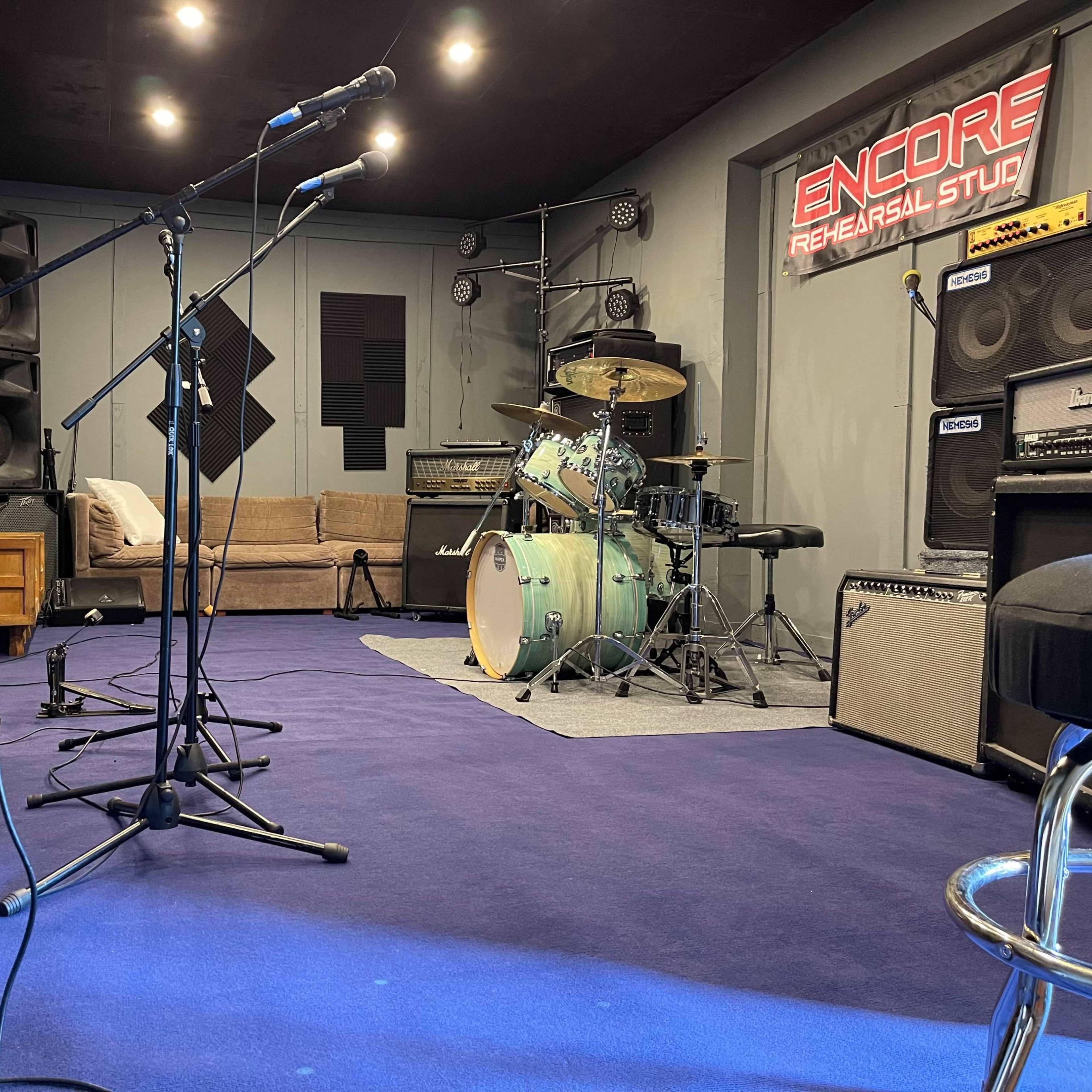 A music rehearsal studio with microphones set up, a drum kit, amplifiers, and a sofa in a soundproofed room.