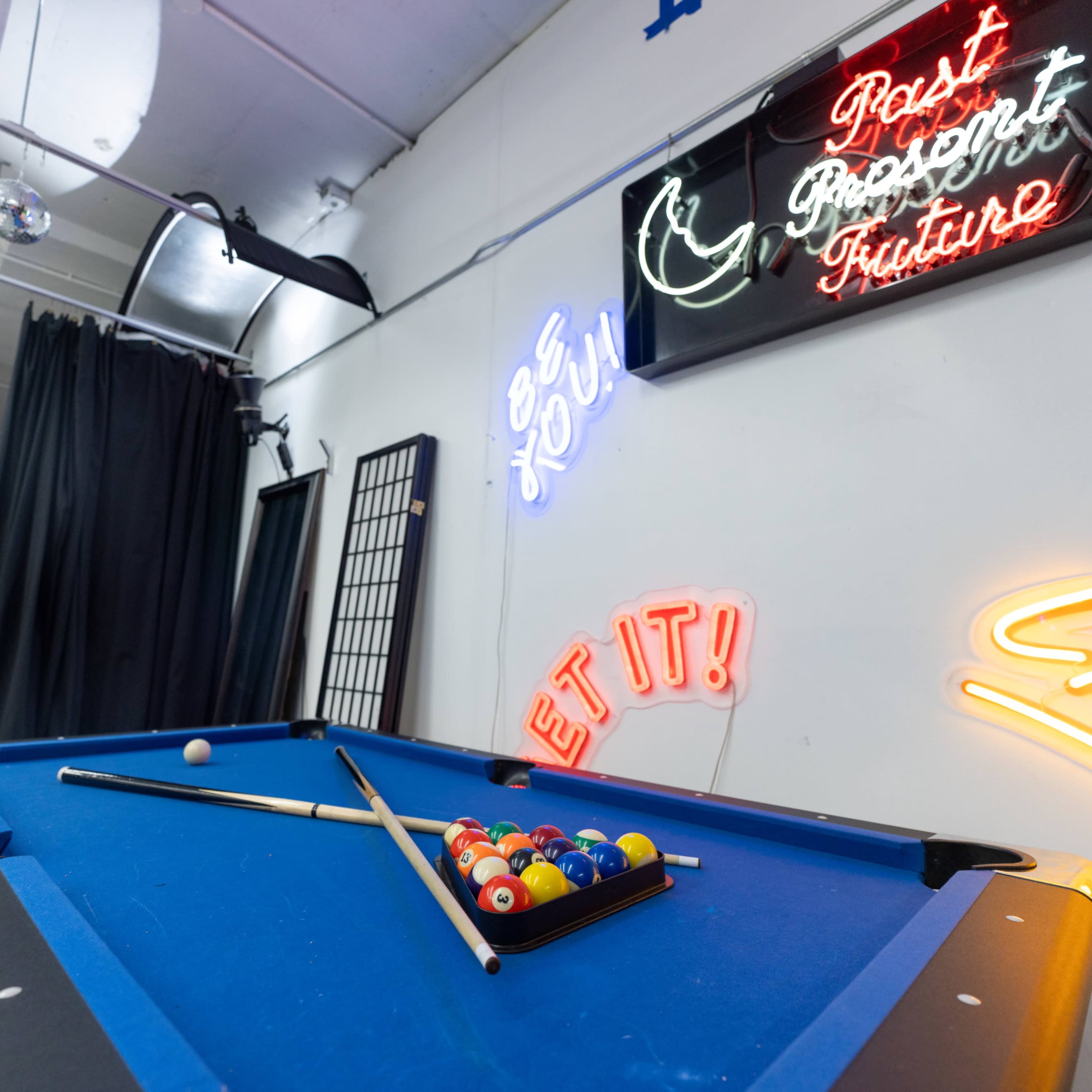 A pool table with balls and cues is positioned beneath colorful neon signs in a well-lit room.
