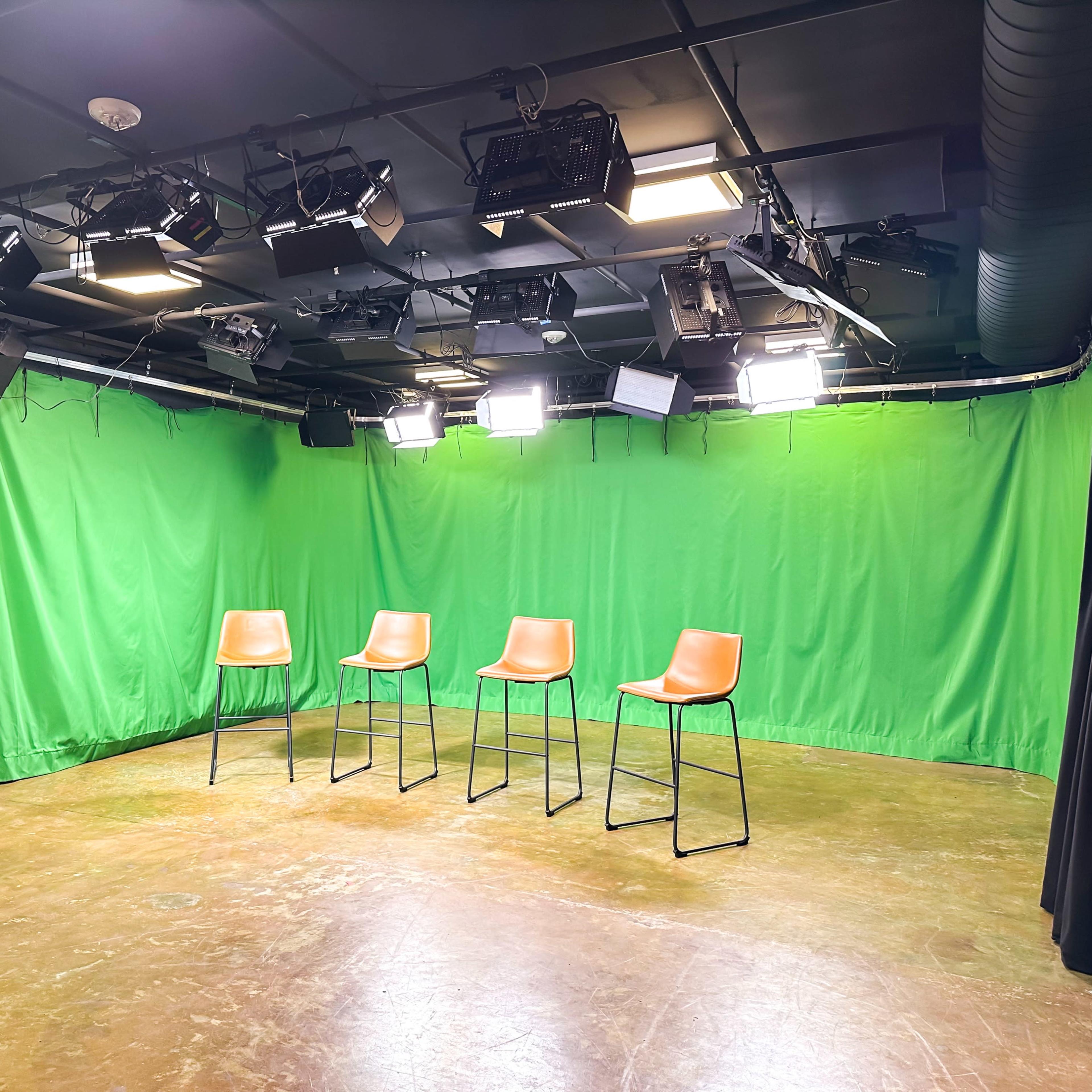 A filming studio with four orange chairs arranged in front of a green backdrop and several overhead lights.