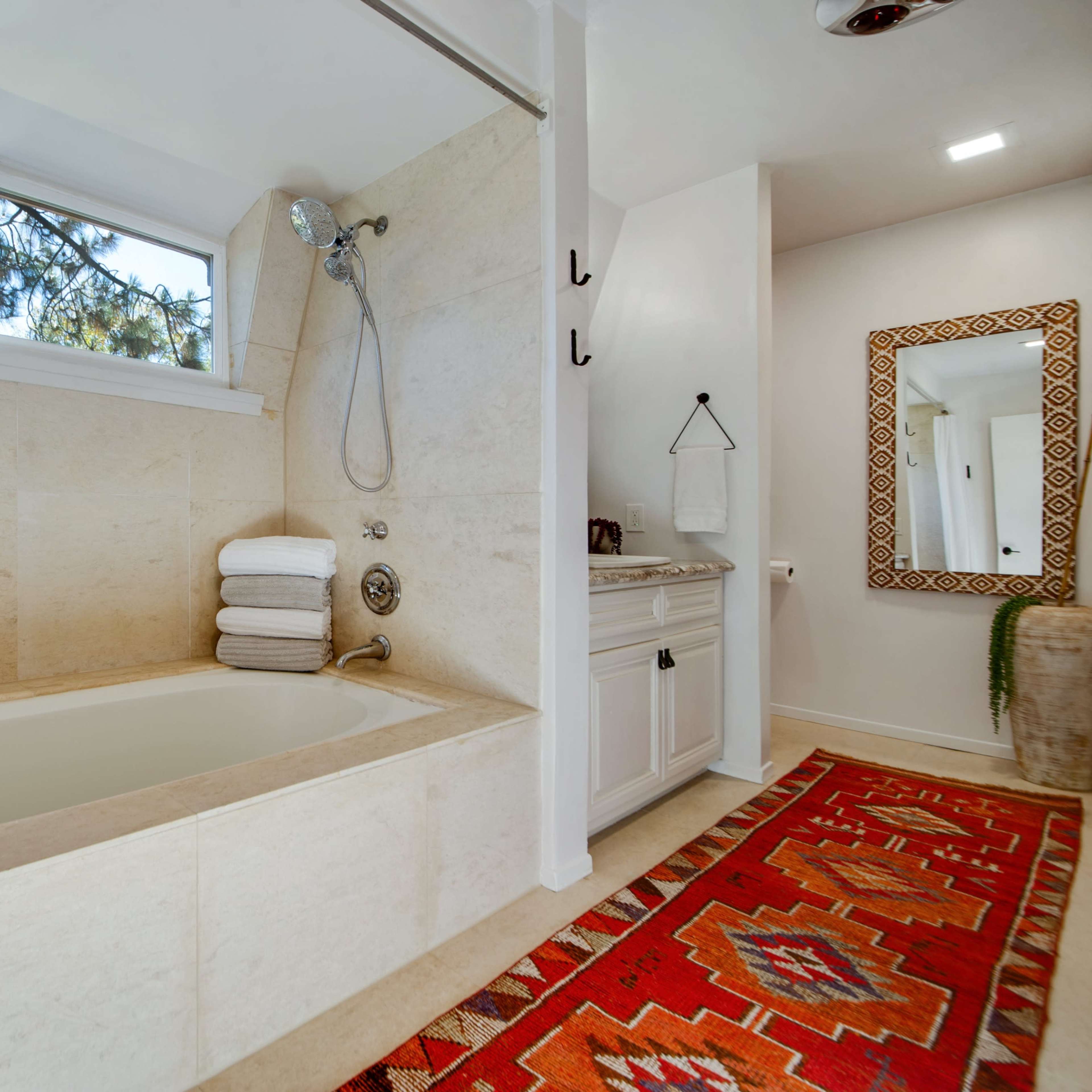 A modern bathroom featuring a beige stone tub, a shower with a handheld showerhead, a towel stack, and a patterned rug in front of the tub.
