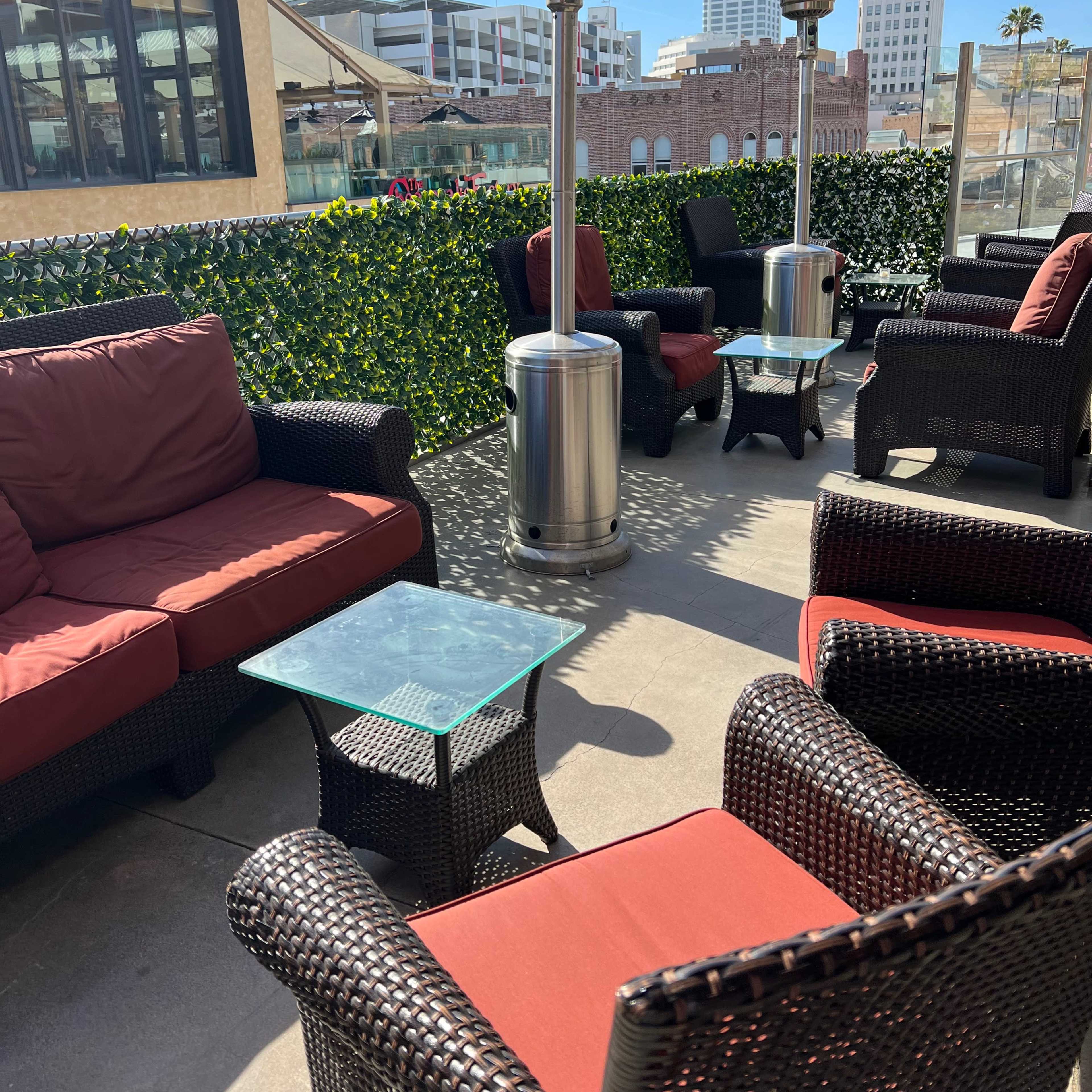 A patio area with seating arrangements that include red cushion chairs, a glass-topped table, and patio heaters surrounded by greenery.