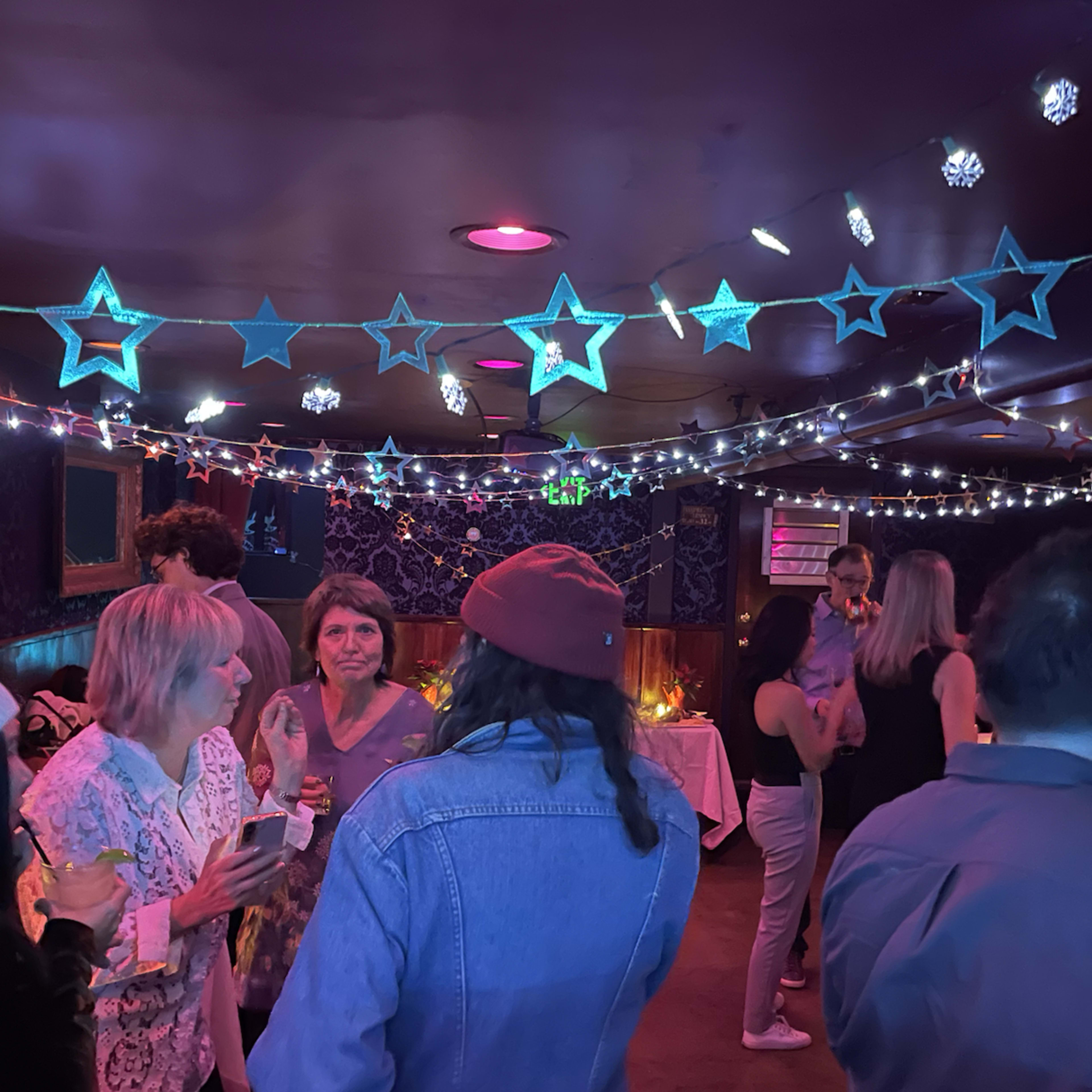 A lively indoor gathering features people socializing under colorful star and string lights in a festive atmosphere.