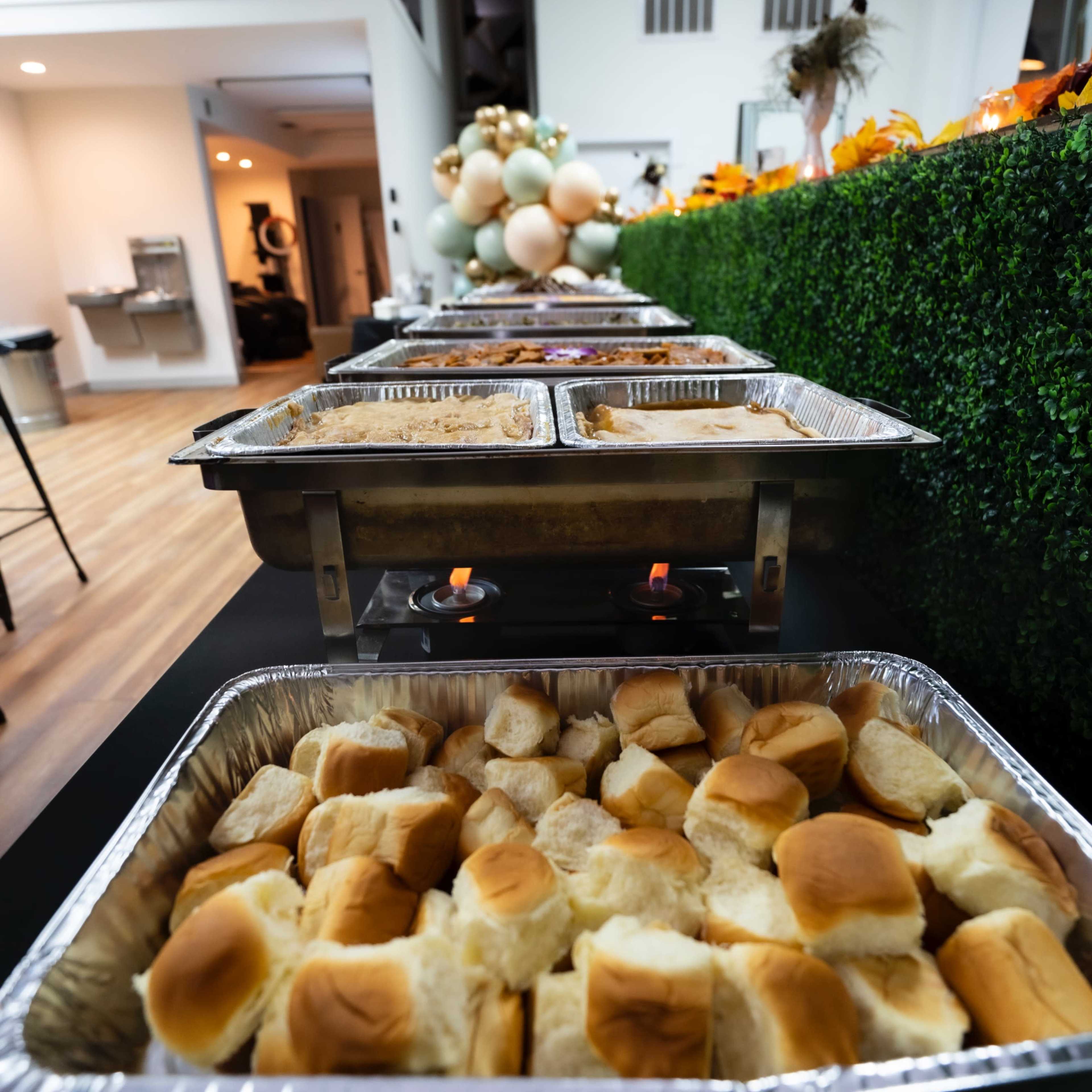 A long table is set up with chafing dishes filled with food and trays of dinner rolls in a well-lit indoor space.