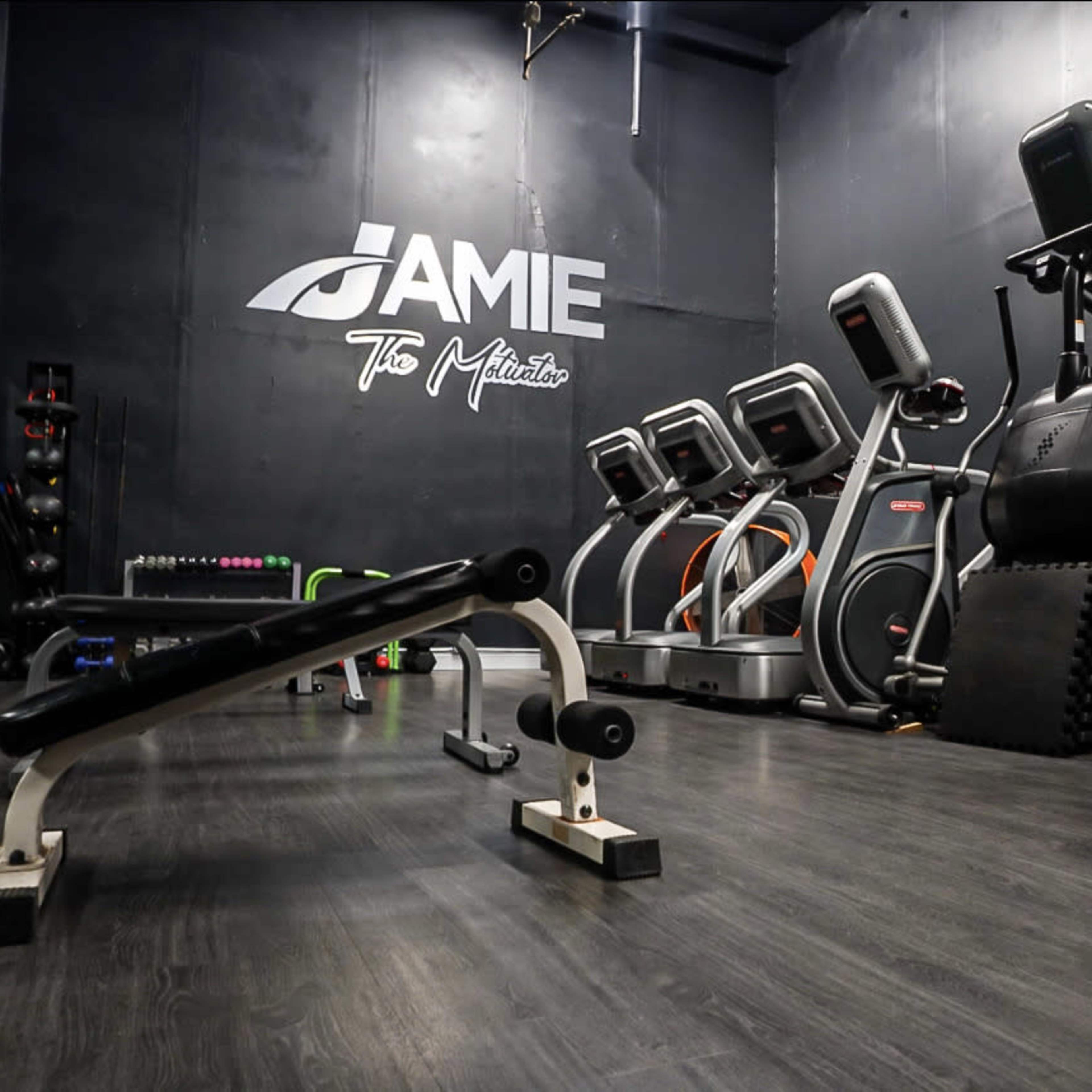 A fitness studio with exercise equipment, including elliptical machines and a weight bench, set against a black wall with the name "Jamie" displayed prominently.