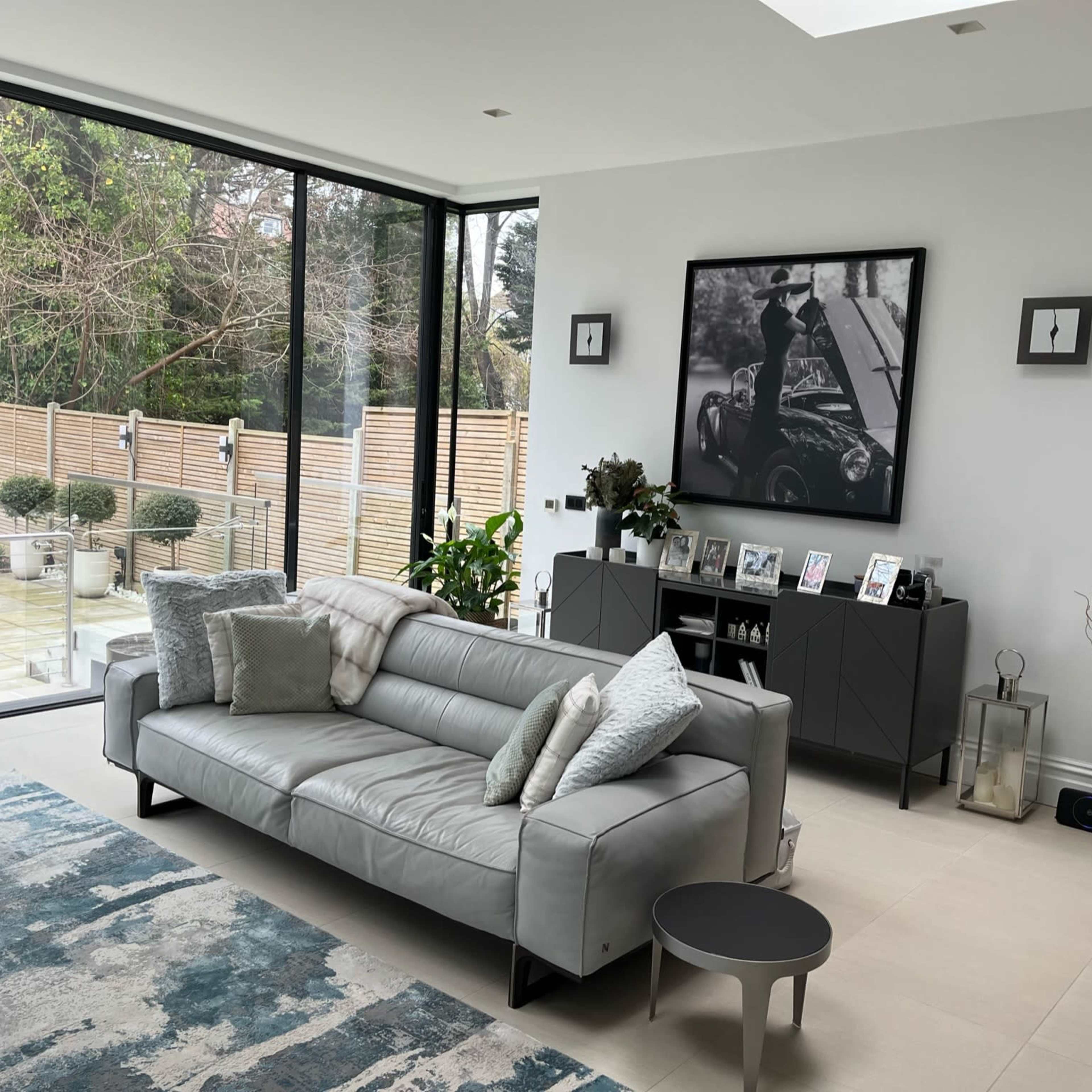 A modern living room with a gray leather sofa, a large black-and-white photograph on the wall, and large windows overlooking a garden.