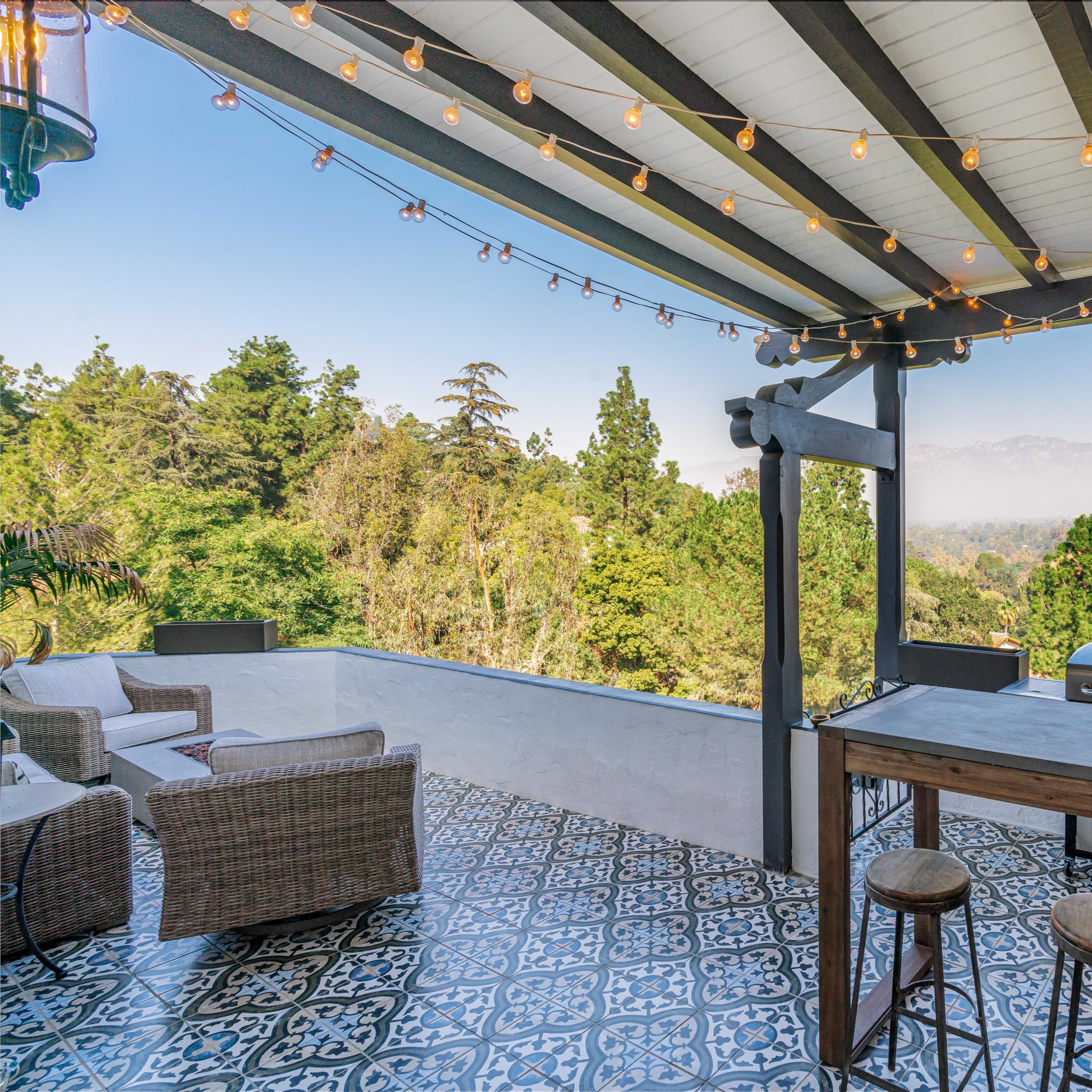 A patio area features a barbecue grill, wooden bar with stools, and string lights, overlooking a lush landscape.