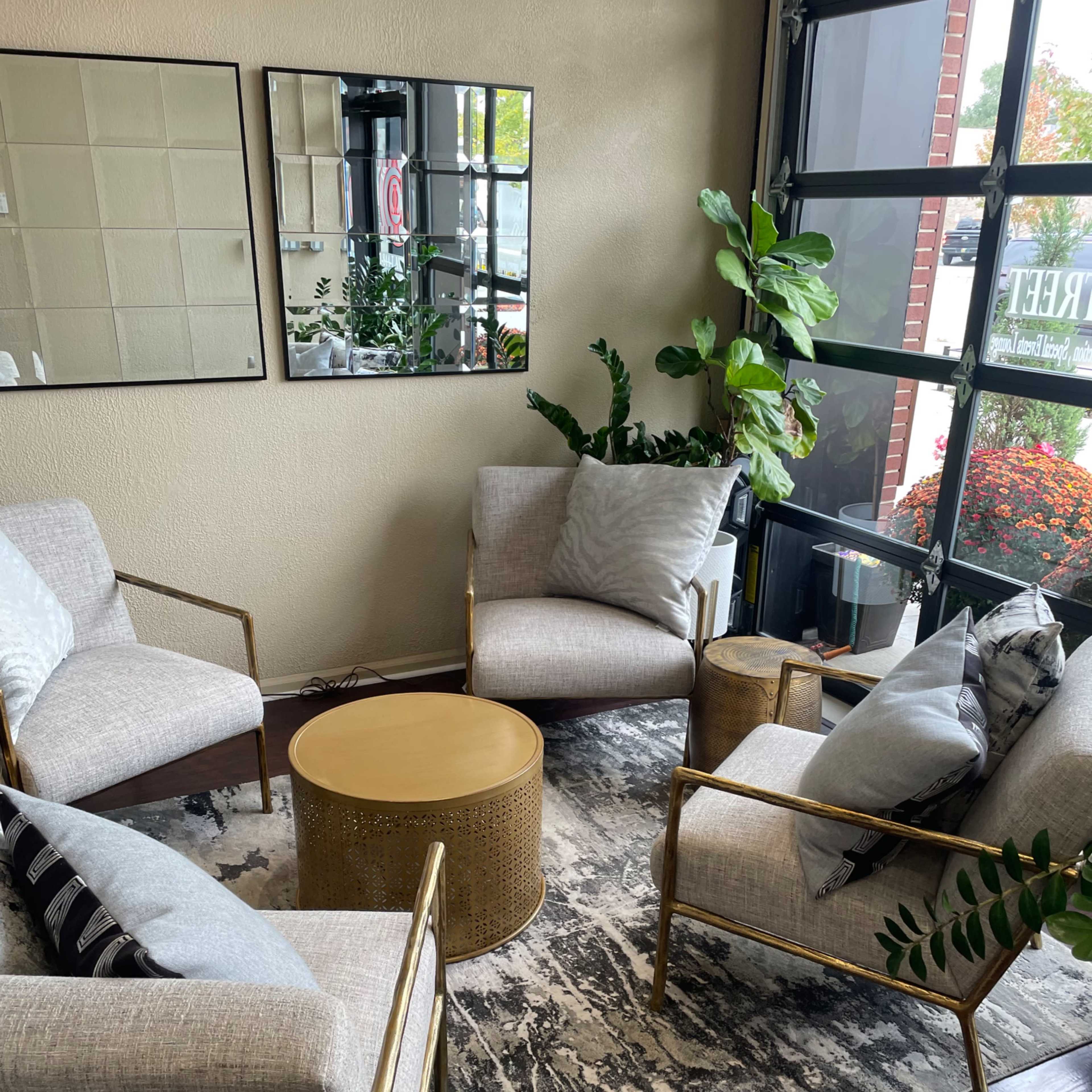 A small seating area with four modern armchairs arranged around a circular gold coffee table on a patterned rug, with large windows and potted plants in the background.