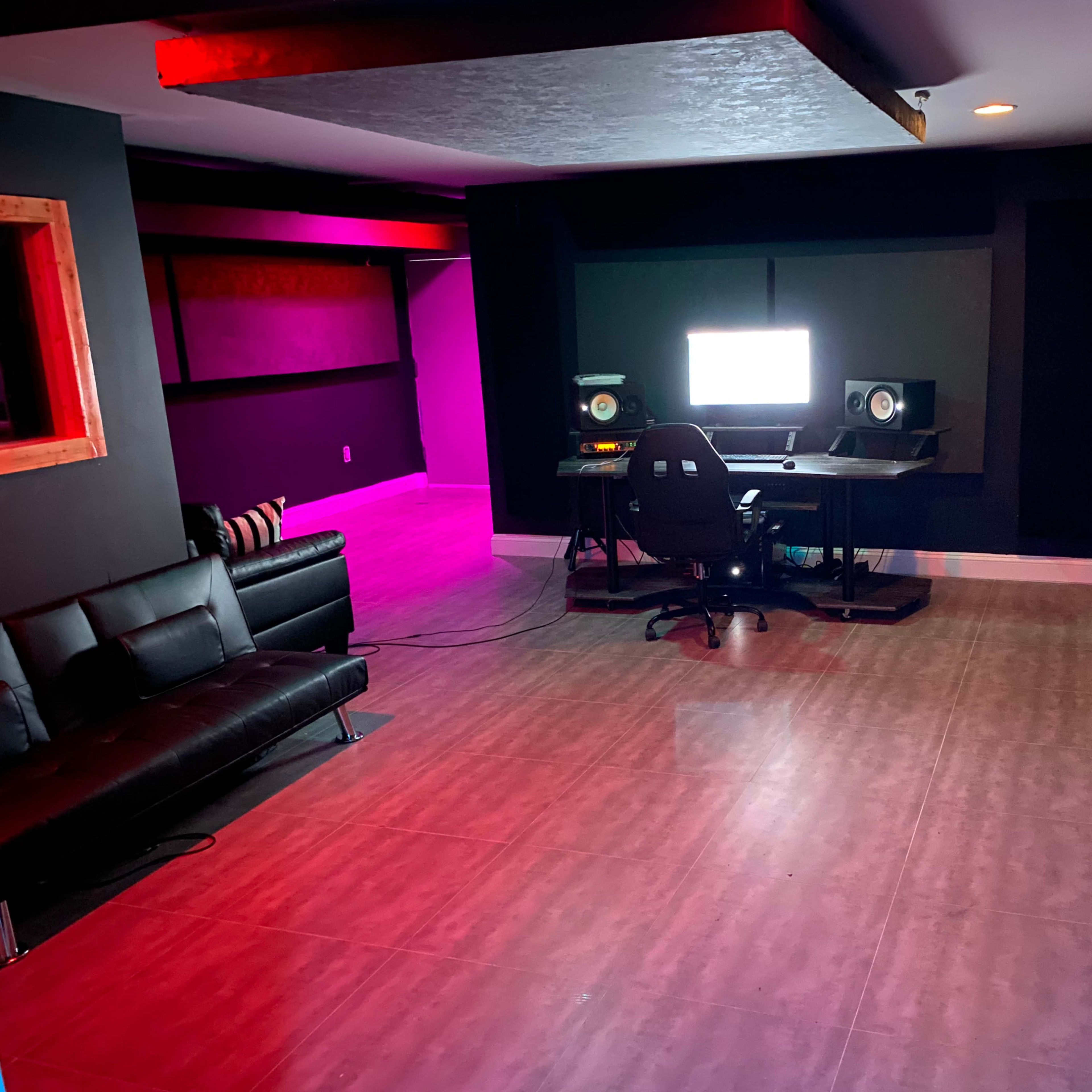 A modern music studio with a computer workstation, speakers, and a black sofa, illuminated by purple and red lighting.