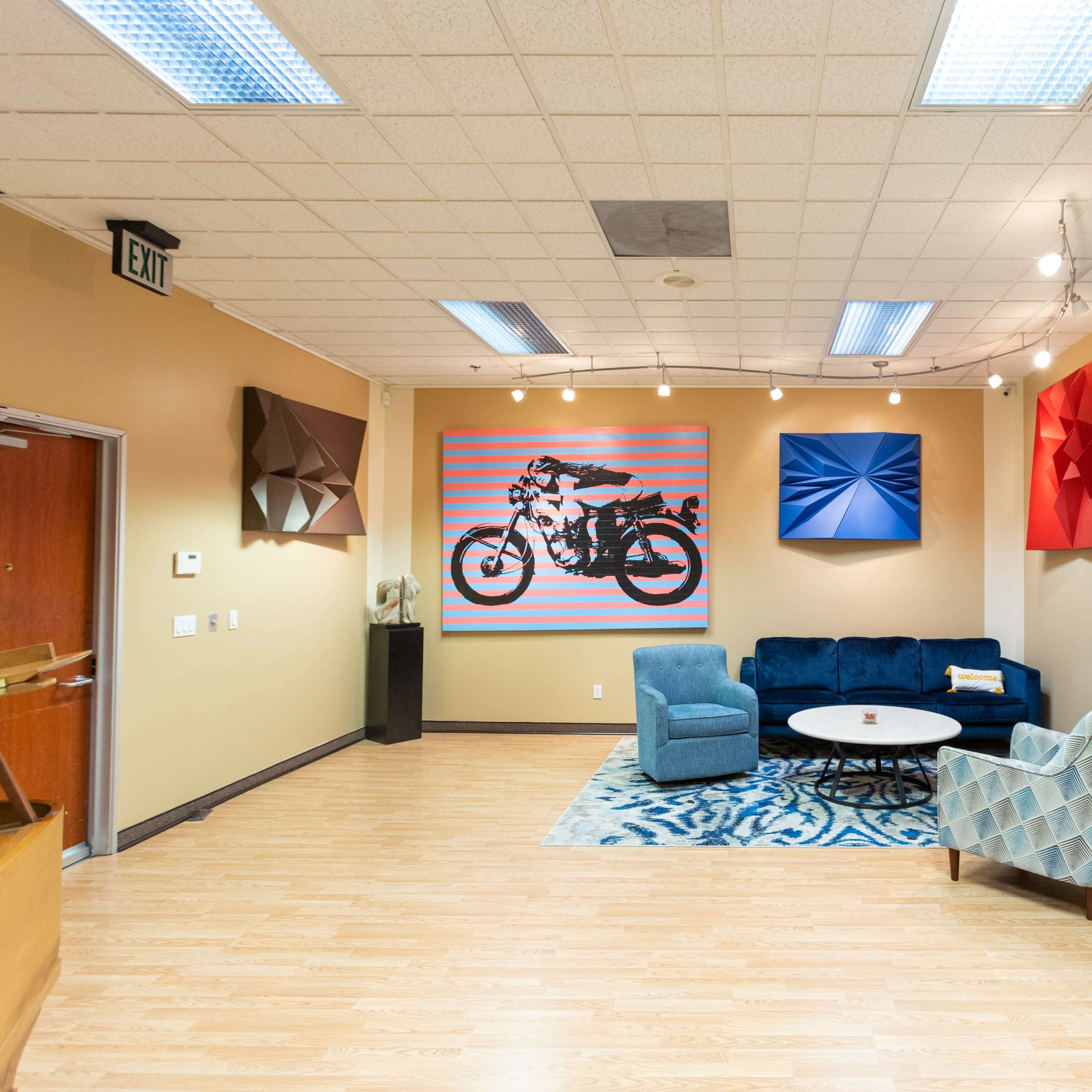 A modern art gallery featuring a blue couch, a circular coffee table, and various colorful artworks on the walls, including a motorcycle and a figure in red.
