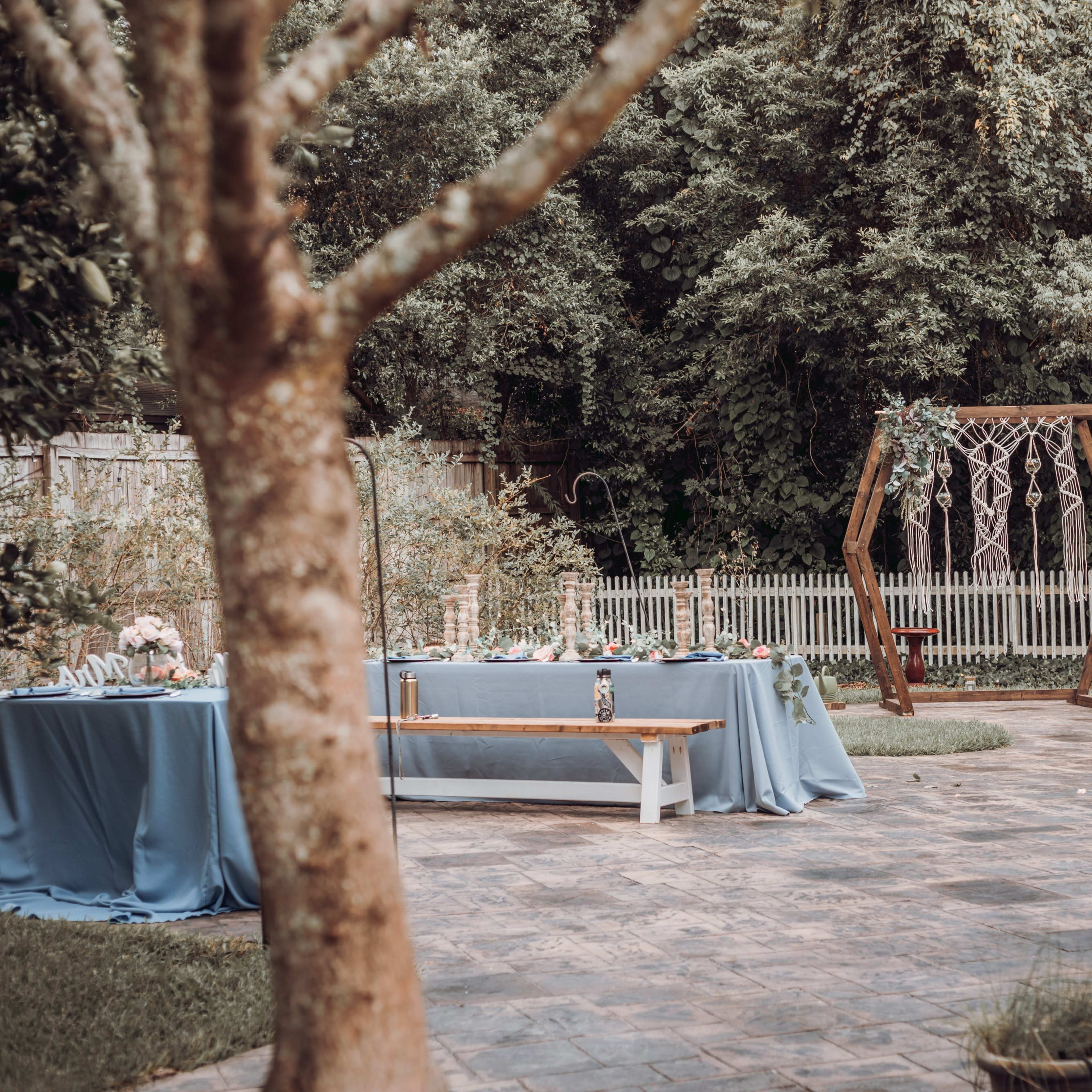 The outdoor area features several tables covered with blue tablecloths, a wooden swing set decorated with greenery, and a stone pathway surrounded by trees.