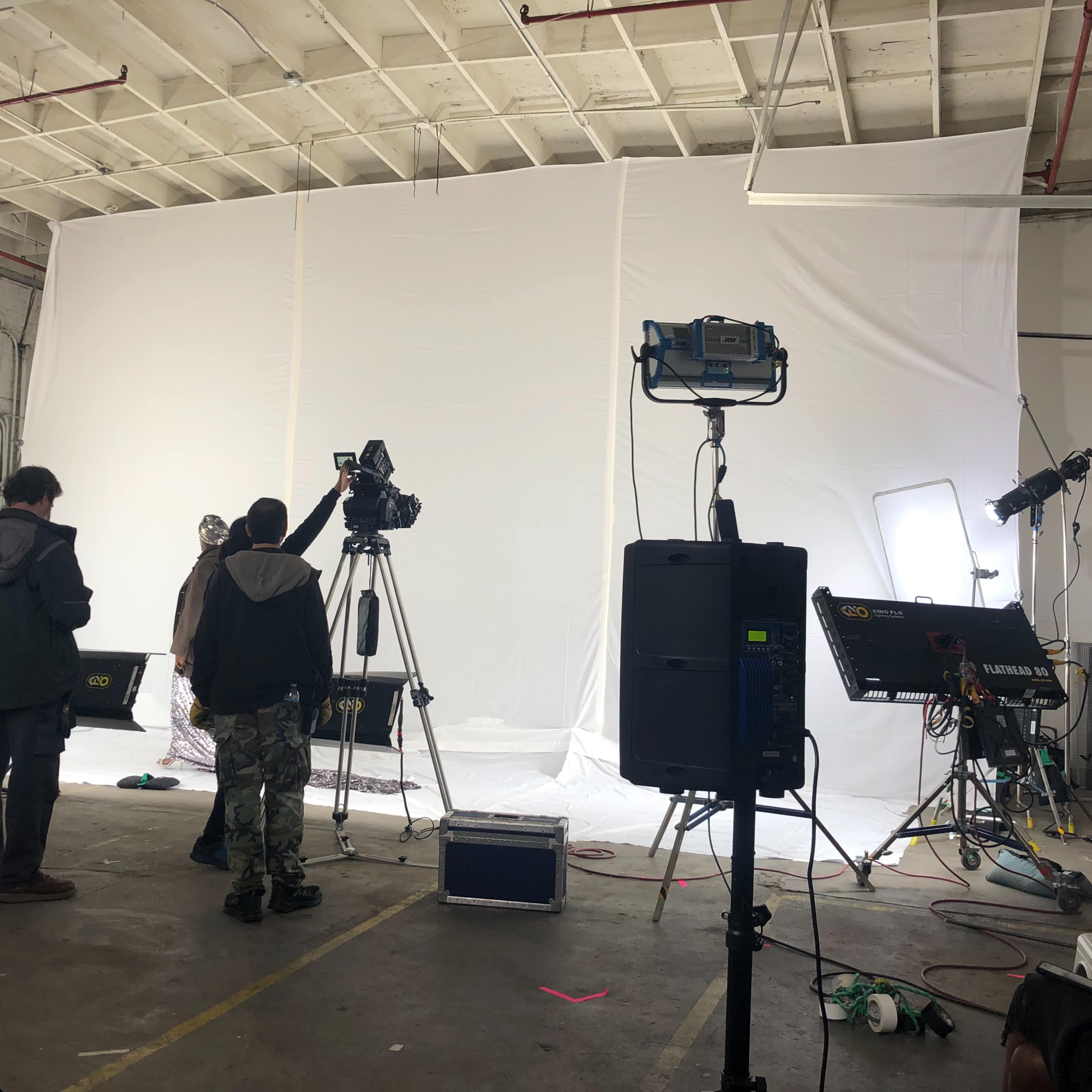 A group of filmmakers is setting up equipment in a studio for a video shoot, with cameras directed at a blank white backdrop.