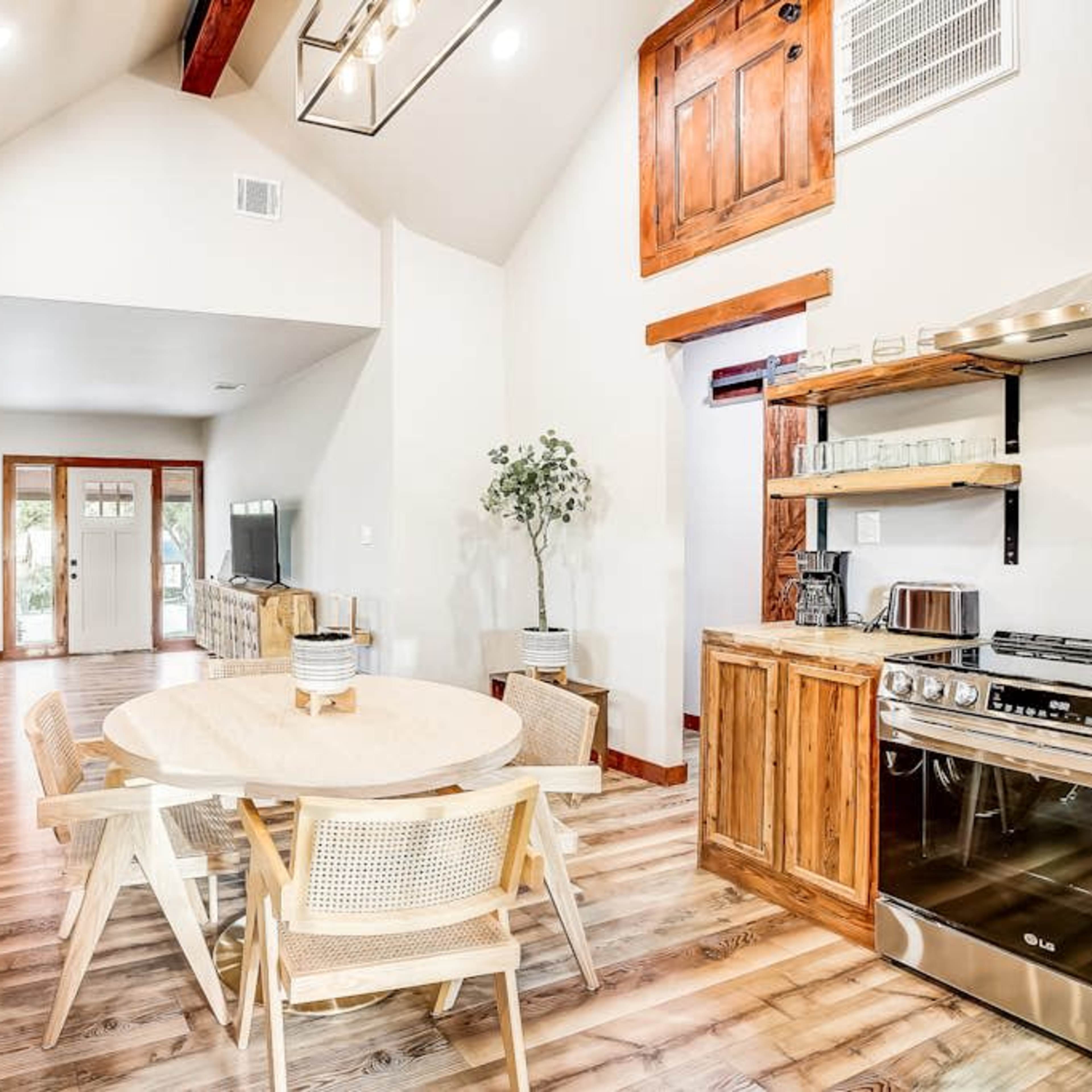 A modern open-concept living space features a round dining table surrounded by light wooden chairs, a rustic kitchen, and large windows allowing natural light to flow in.