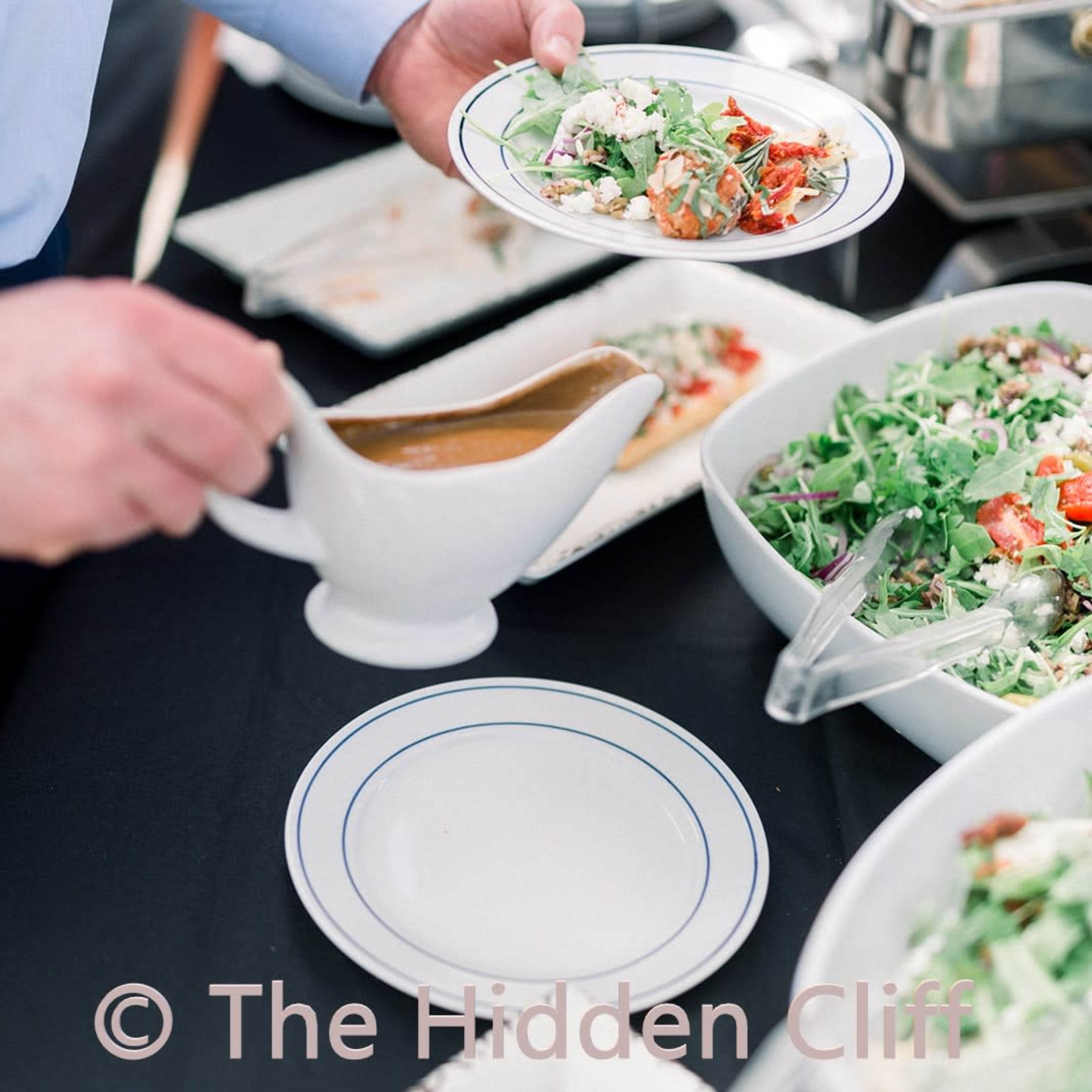 A person is serving food from a buffet, holding a plate while preparing to add sauce from a gravy boat.