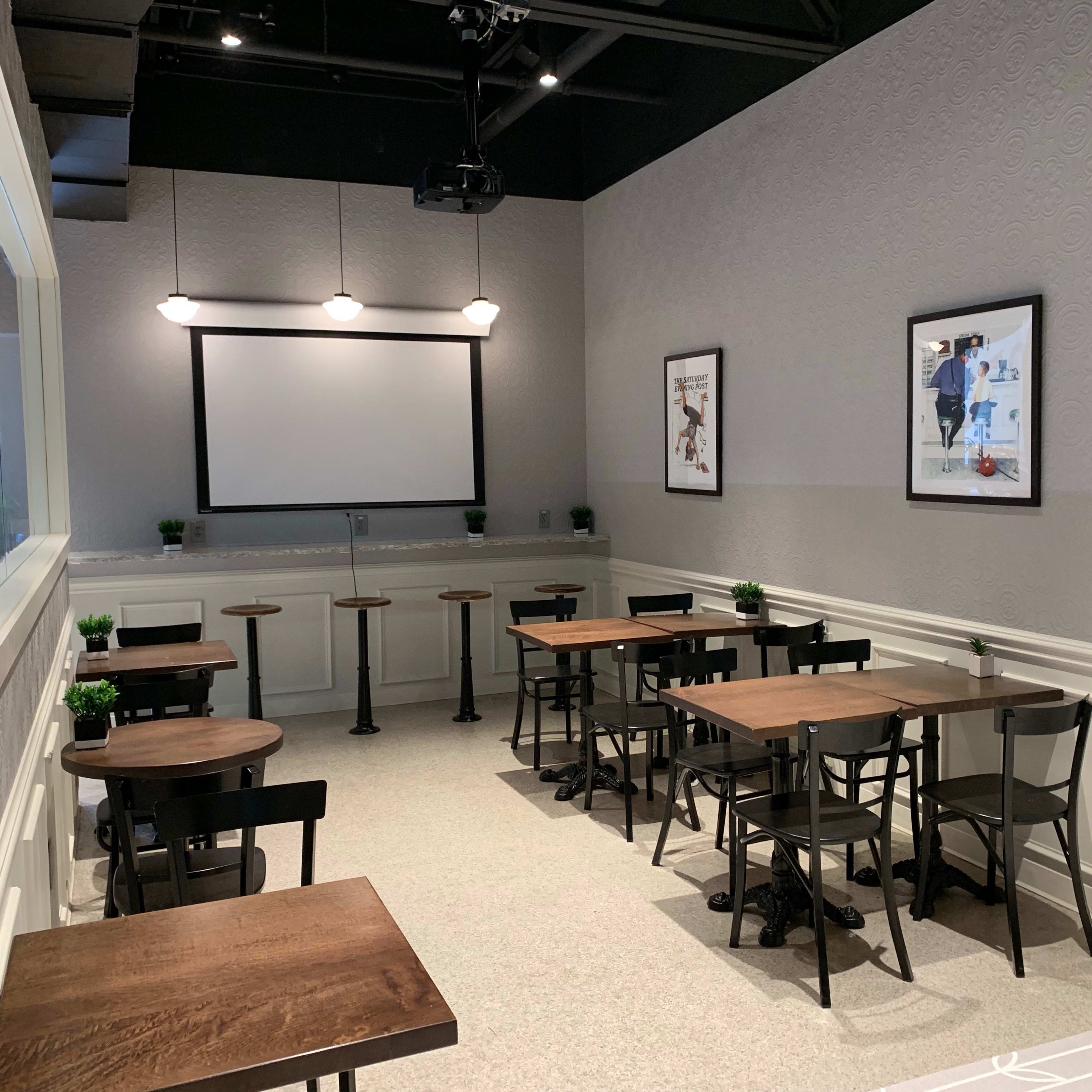 A modern café interior with several tables and stools arranged against a light gray wall featuring a projector screen and framed artwork.
