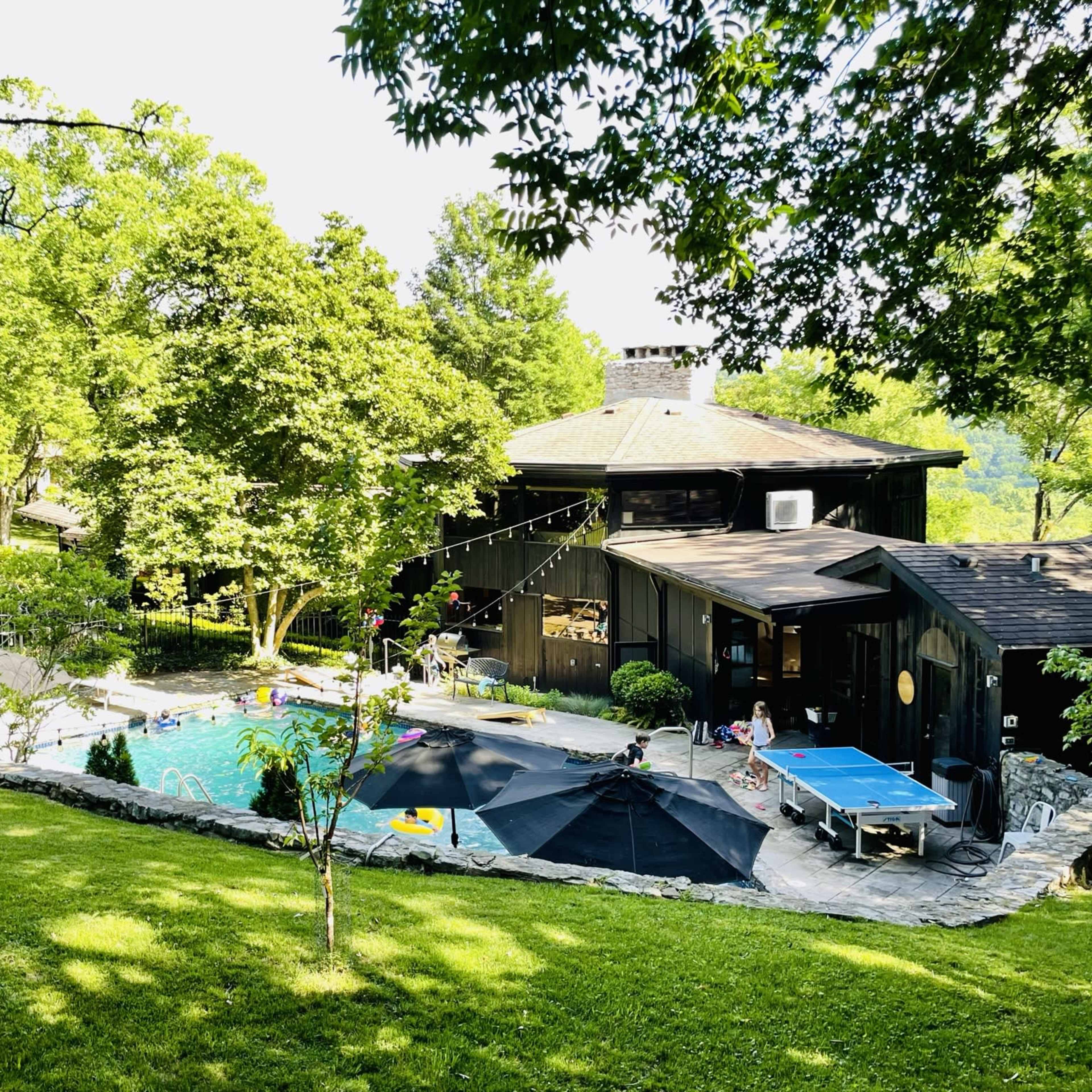 A wooded backyard features a black house with a pool surrounded by lounge areas and outdoor games.