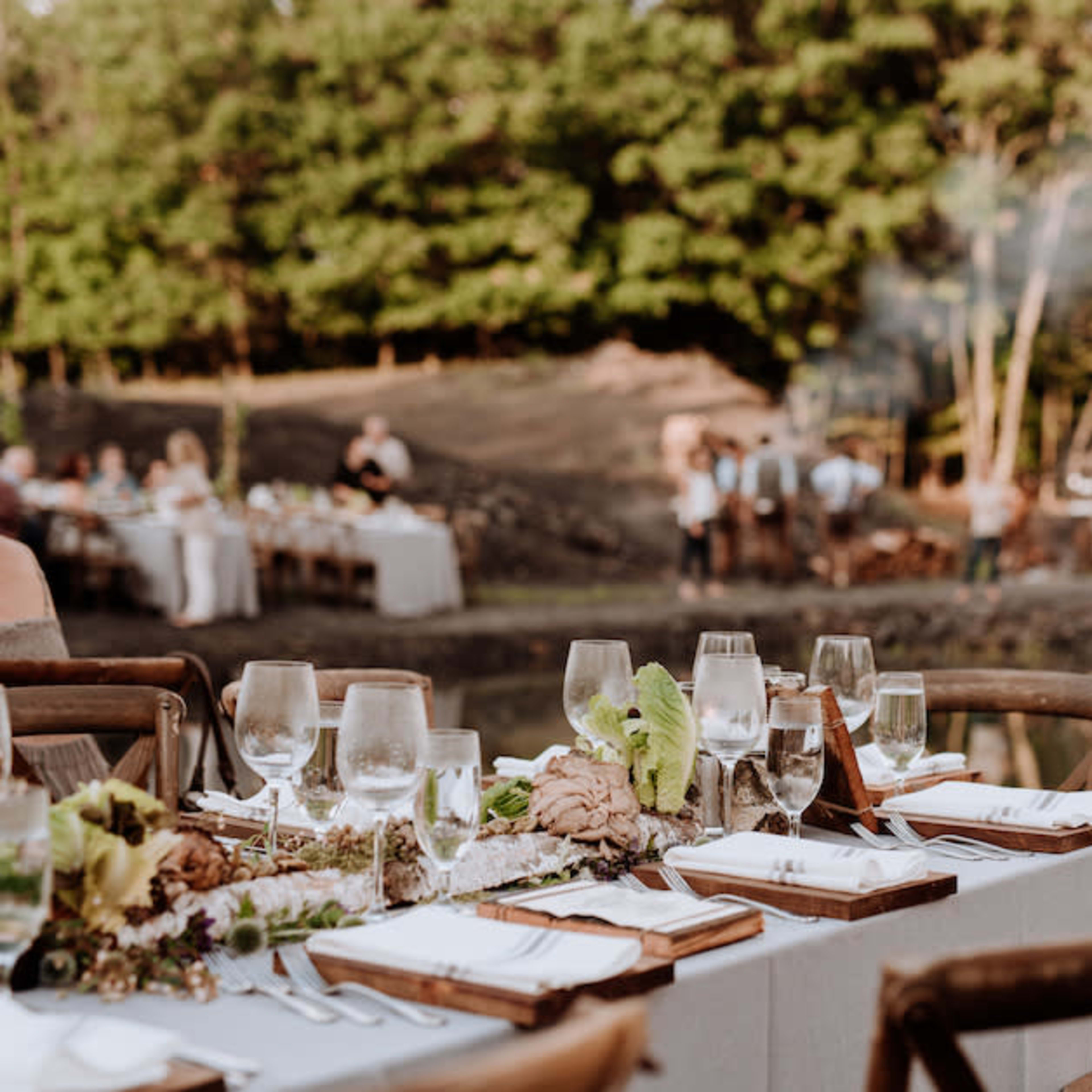 A long table set with glasses and tableware is arranged near a scenic outdoor setting with greenery and a small body of water in the background, where guests are gathered for an event.