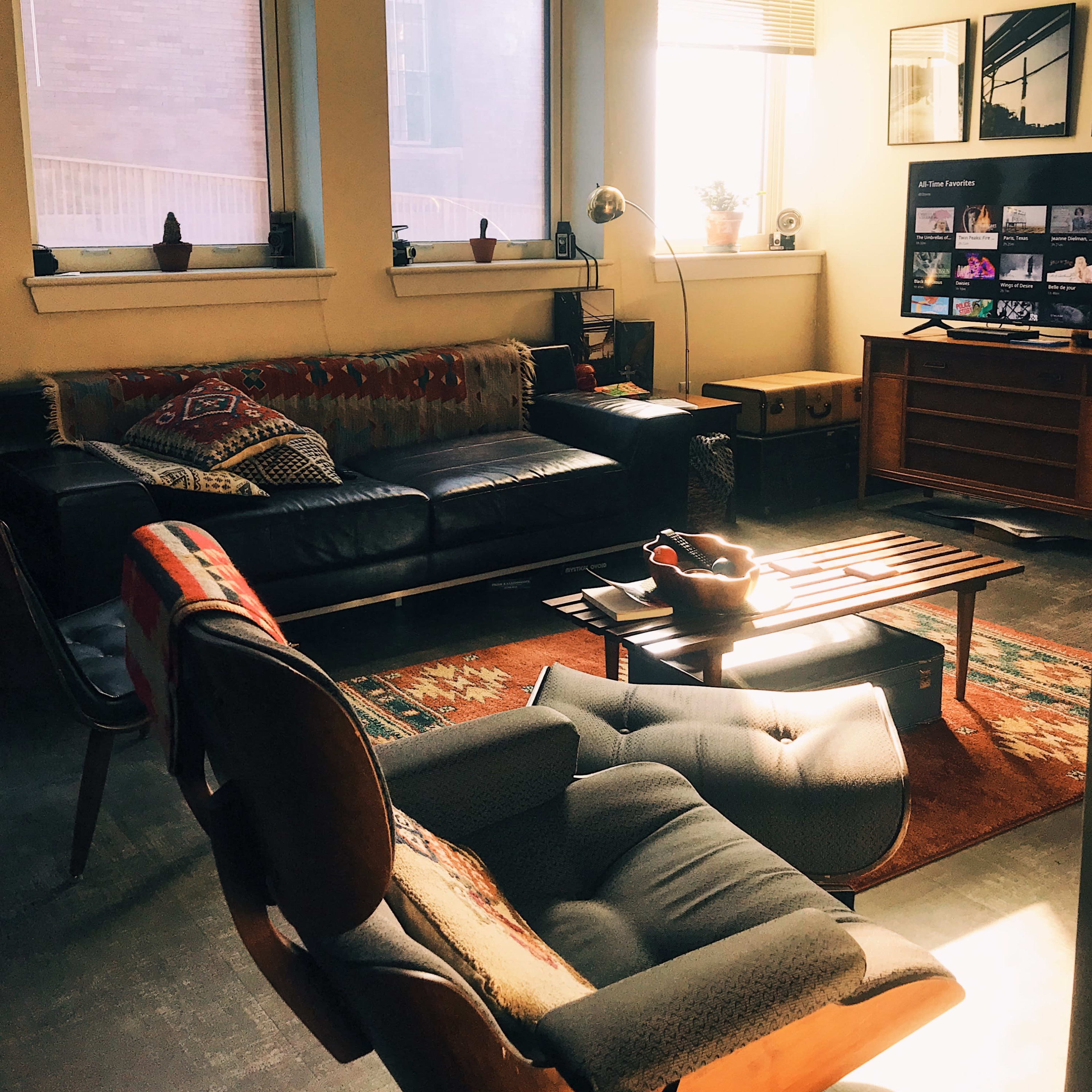 The living room features a leather sofa, a wooden coffee table, an armchair, and large windows allowing natural light to stream in.