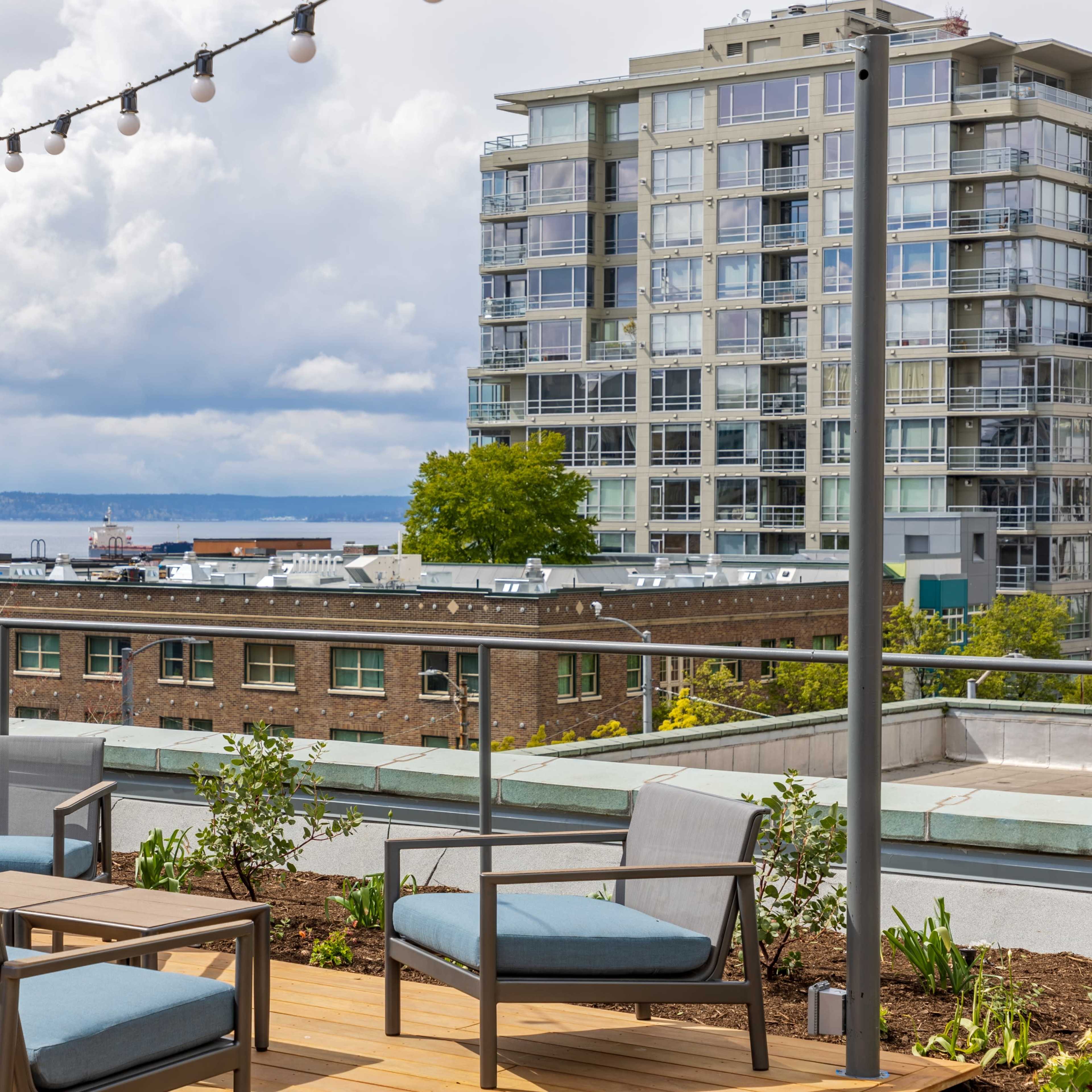 A rooftop patio features modern furniture and a view of a waterfront city skyline under cloudy skies.