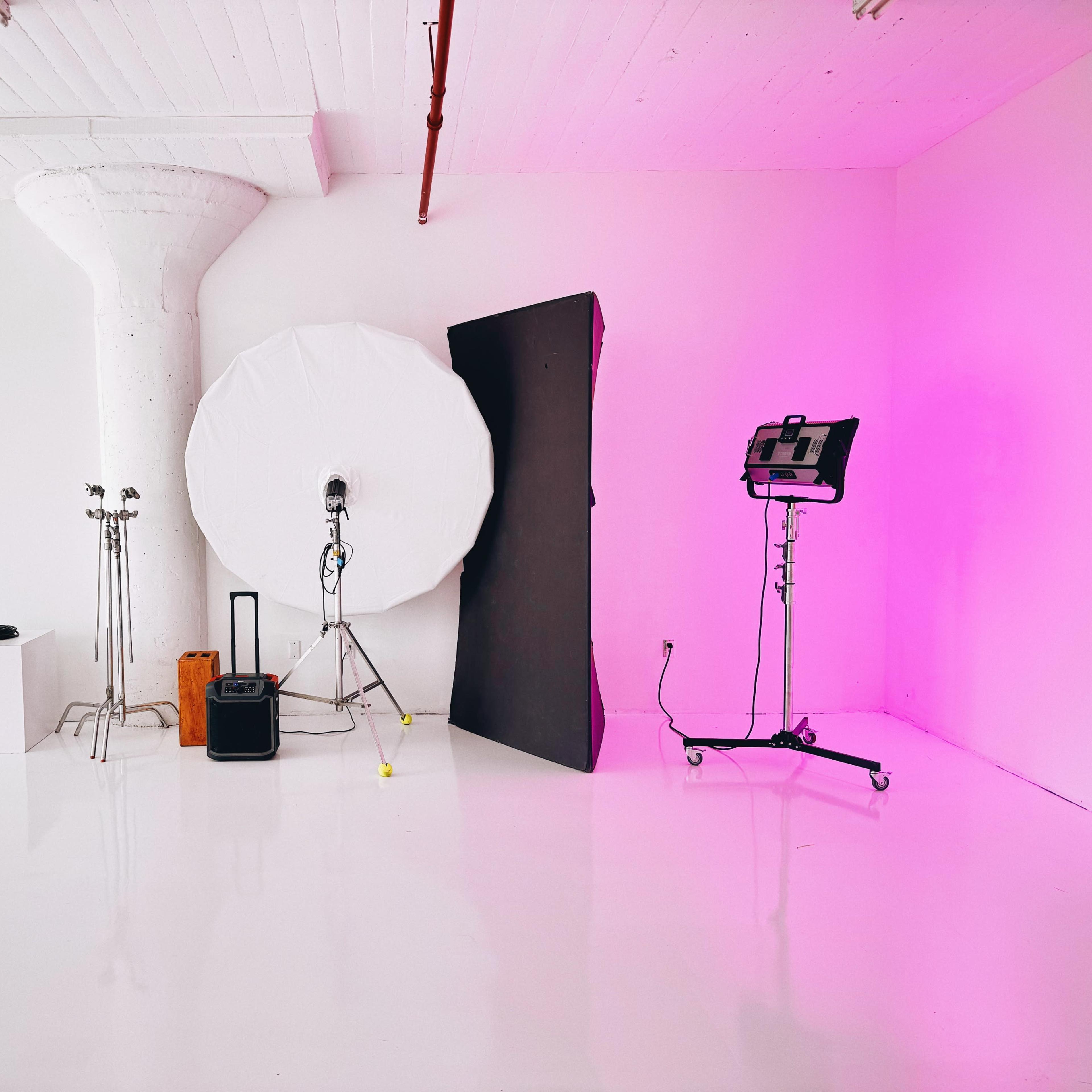 A minimalist studio setup with lighting equipment, a large circular reflector, and a plain white backdrop against a bright, white floor.