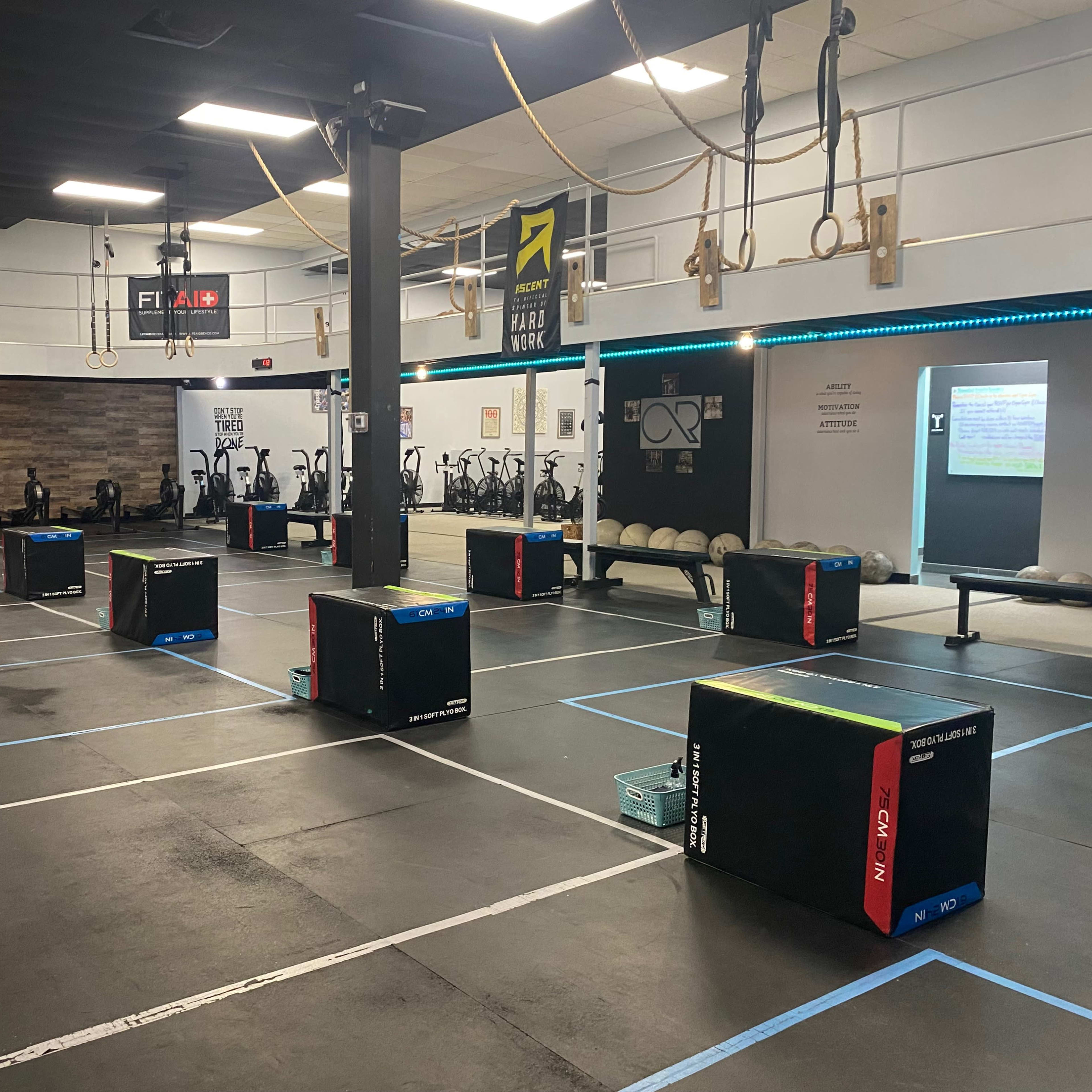An indoor gym with marked workout areas, black boxes arranged in each section, and exercise equipment in the background.
