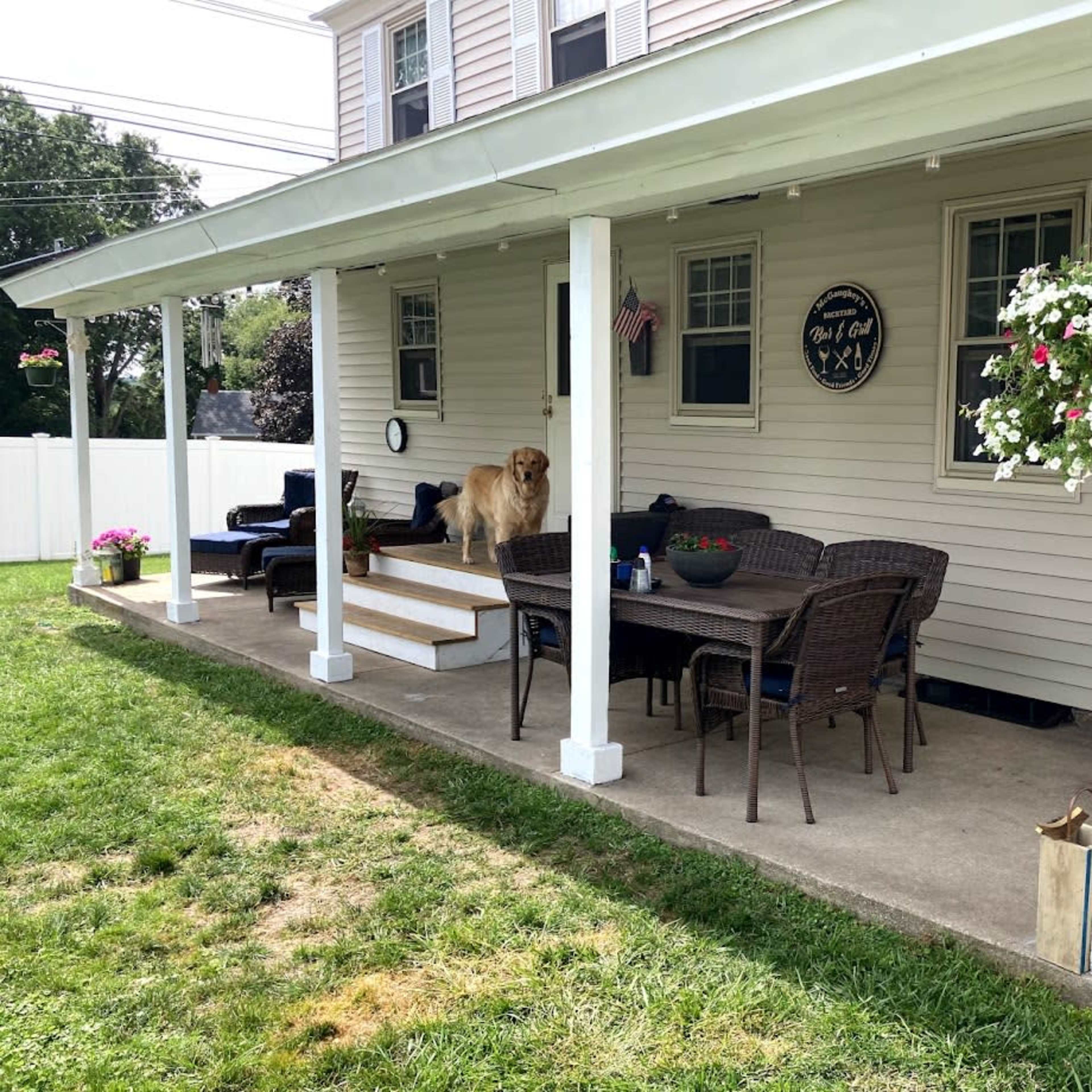A covered patio with a dining table, chairs, a golden retriever, and hanging flower pots.
