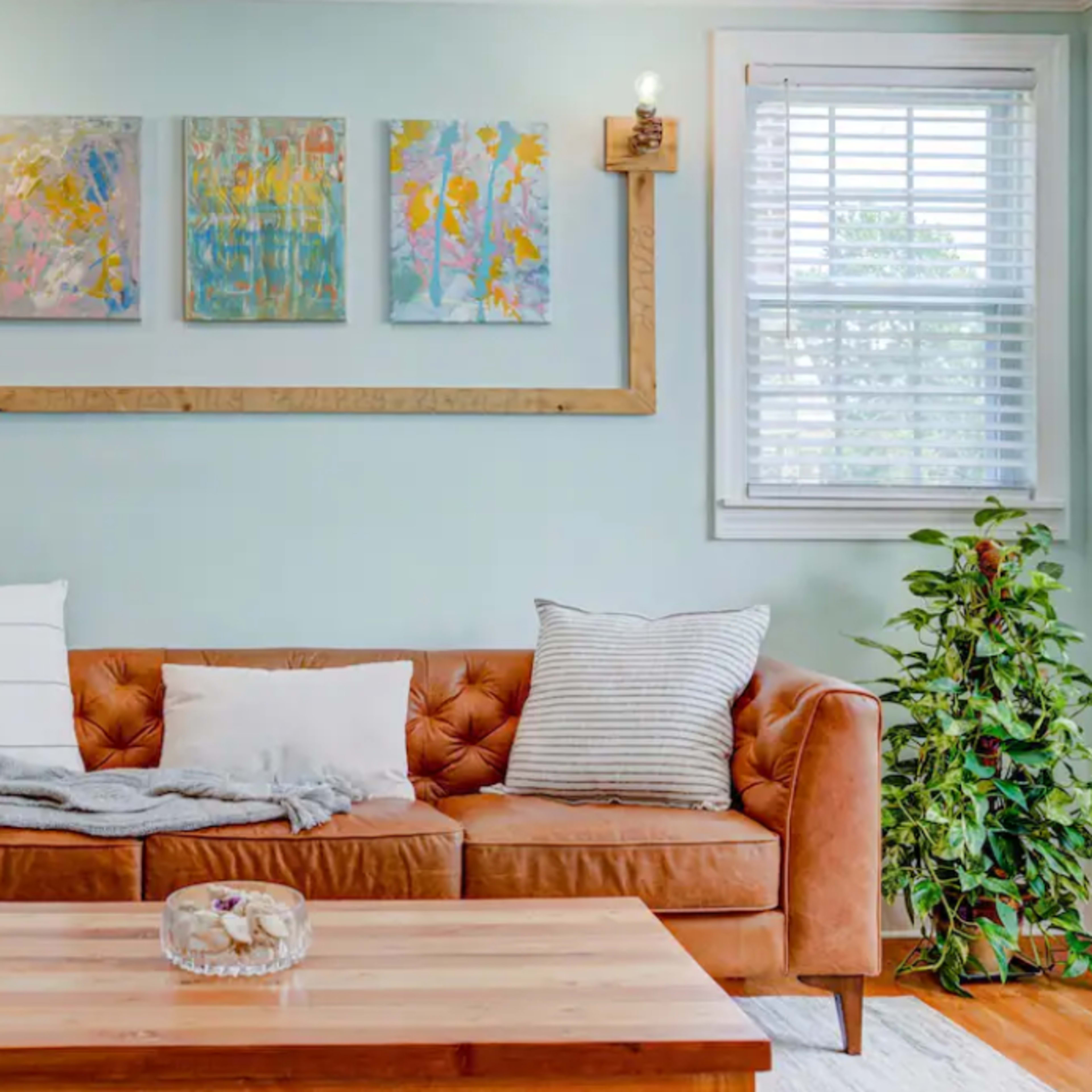 A brown leather sofa with decorative pillows is positioned in front of a light wooden coffee table, against a mint green wall adorned with three colorful paintings and flanked by windows.