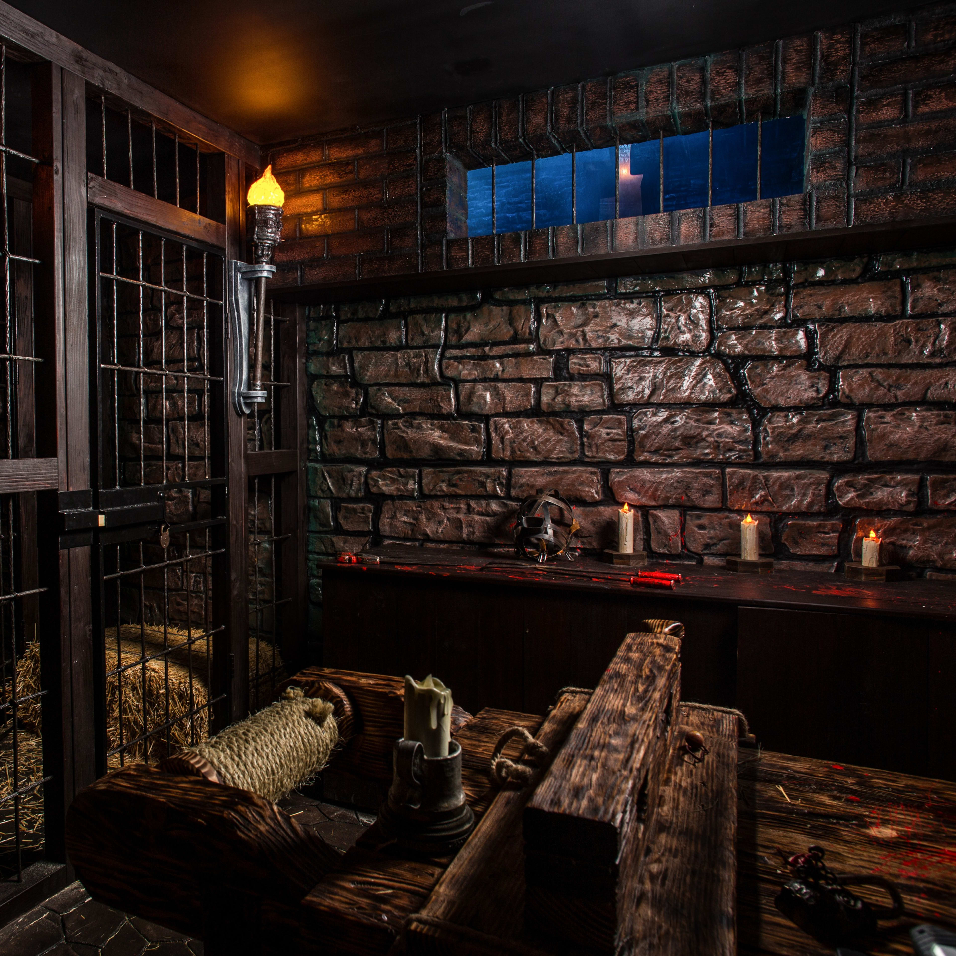 A dimly lit dungeon with stone walls, a barred cage, and a wooden stocks contraption, featuring scattered hay and candles.