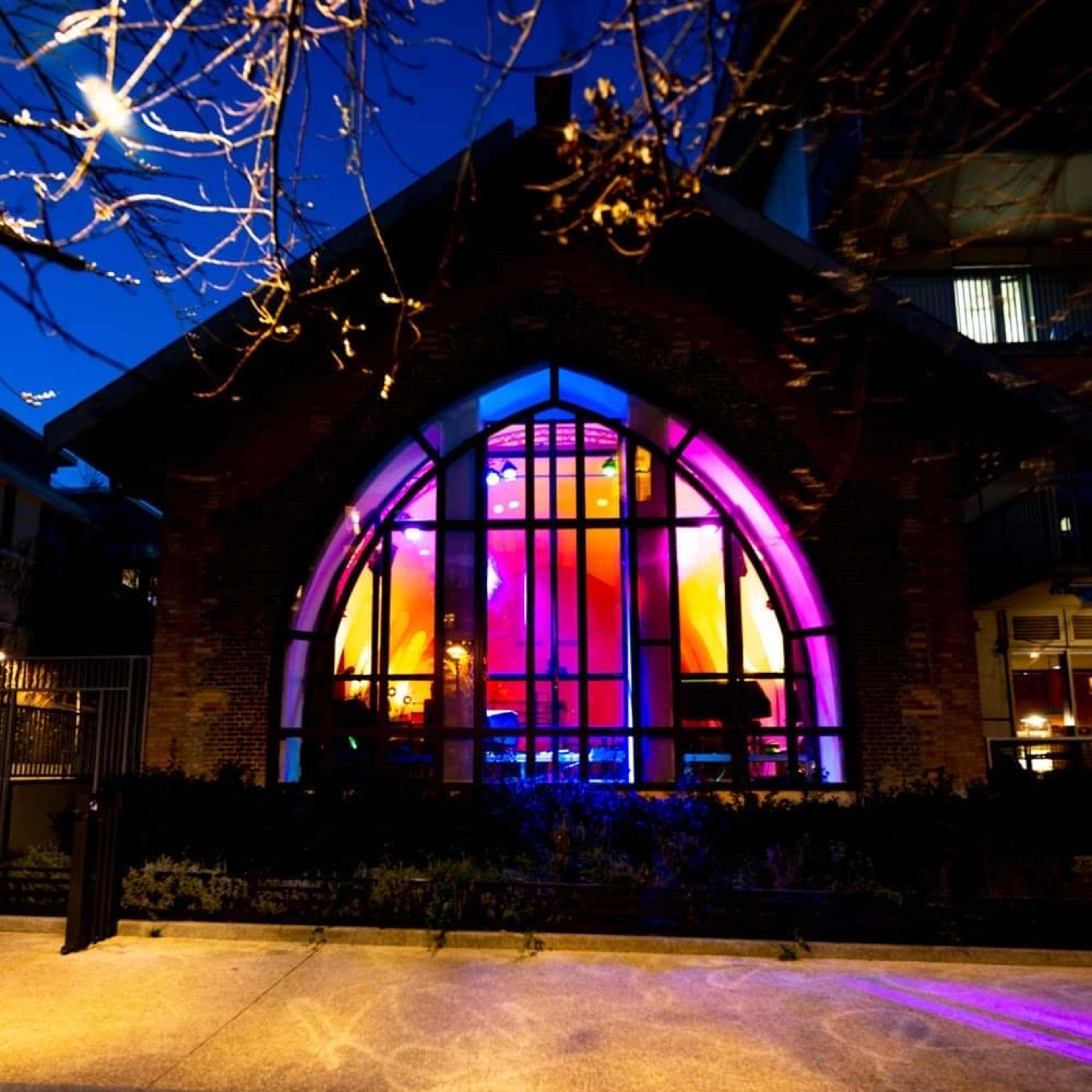 A brick building with an arched window illuminated in vibrant colors against a twilight sky.