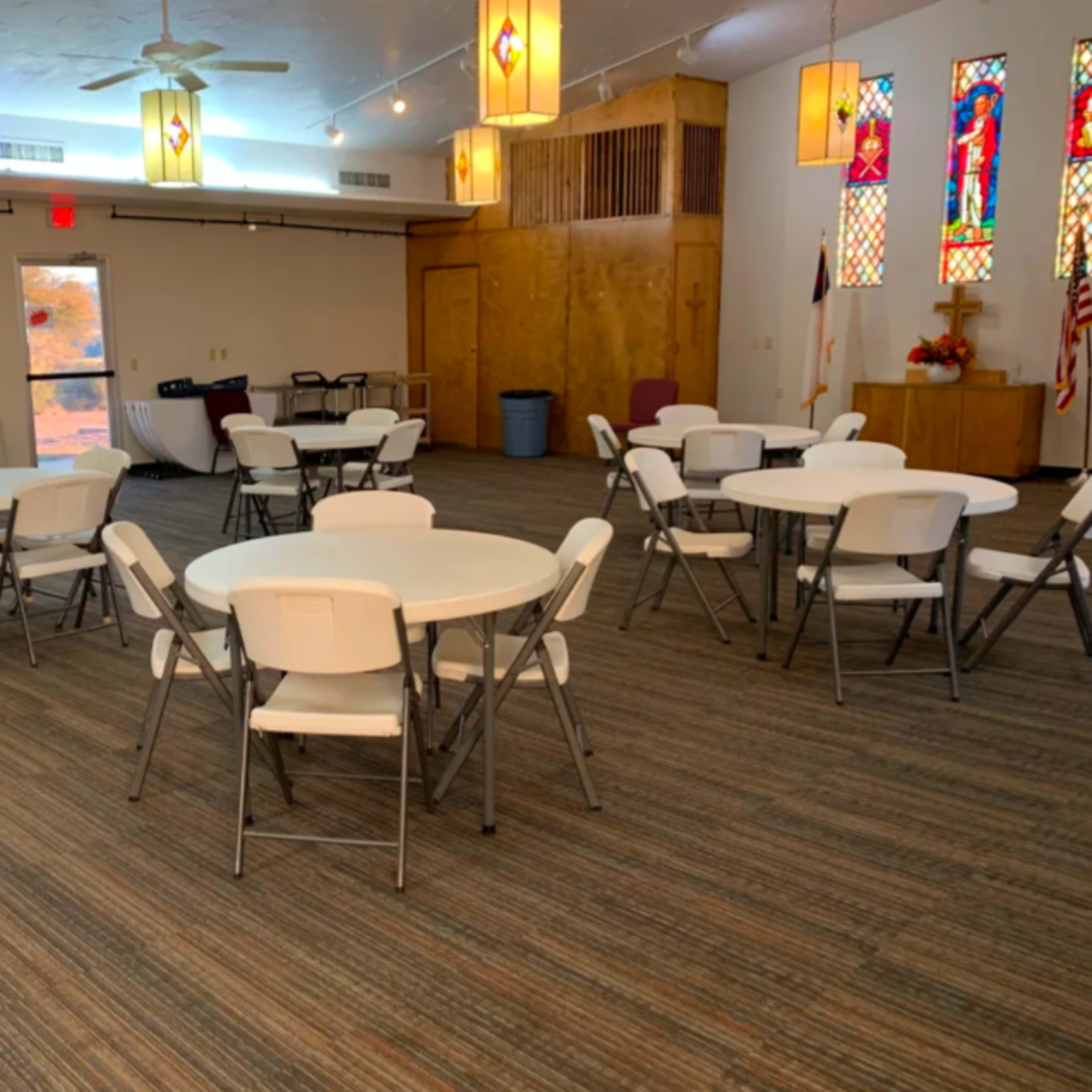 A spacious room with several round tables and chairs arranged for an event, featuring stained glass windows and a wooden stage.