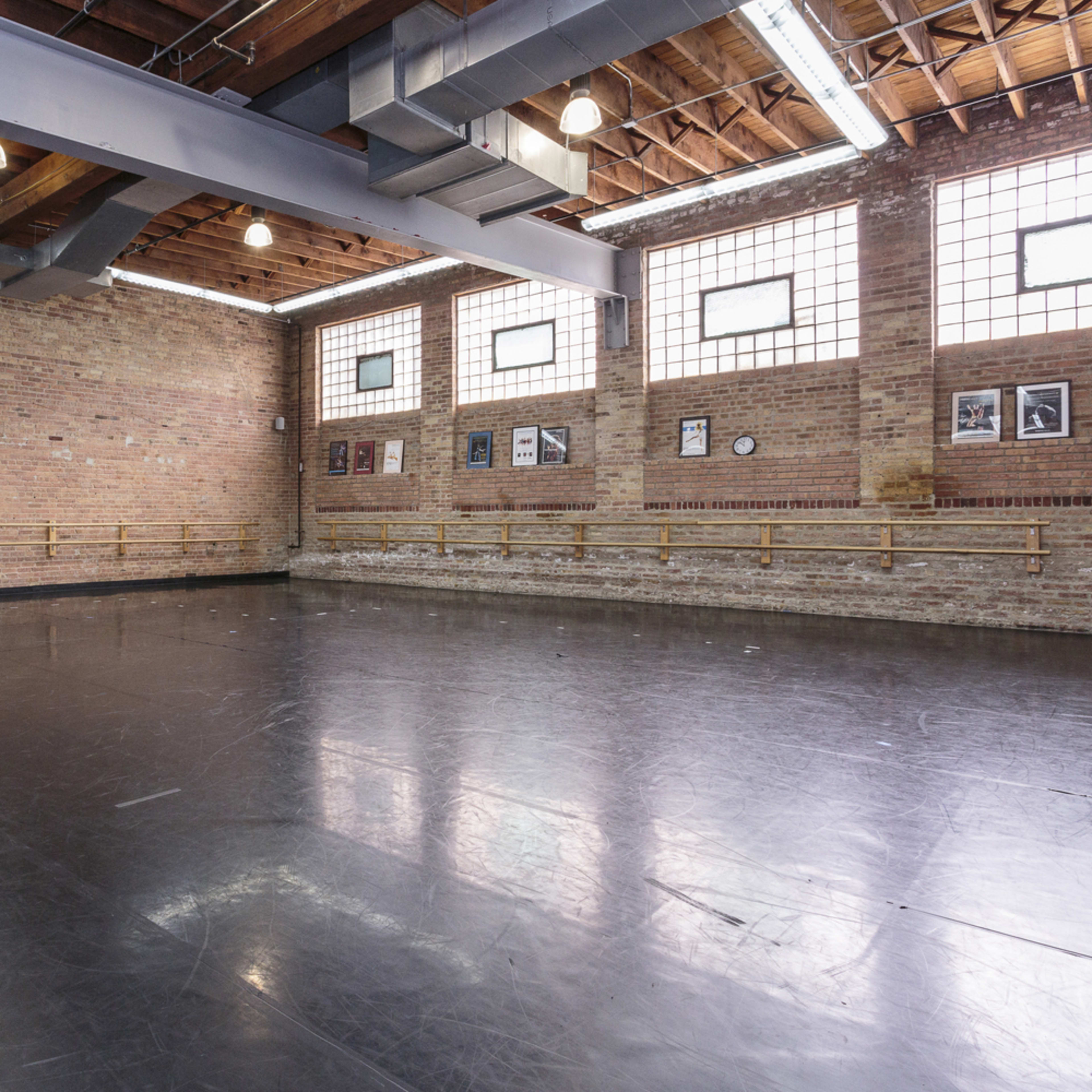 A spacious dance studio features a black flooring surface, exposed brick walls, and large windows allowing natural light to enter.