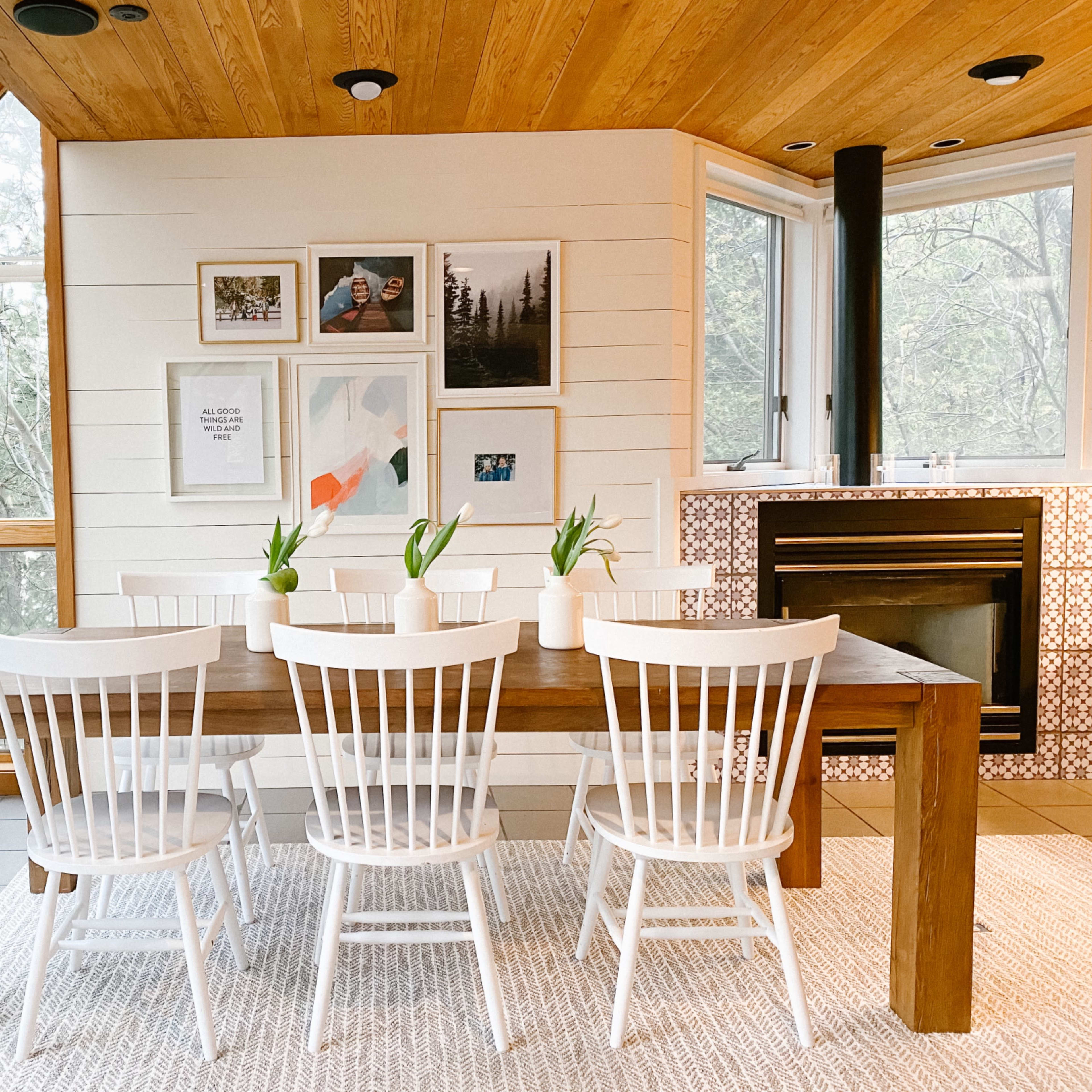 A wooden dining table surrounded by white chairs is positioned in a room with large windows and framed artwork on the walls.