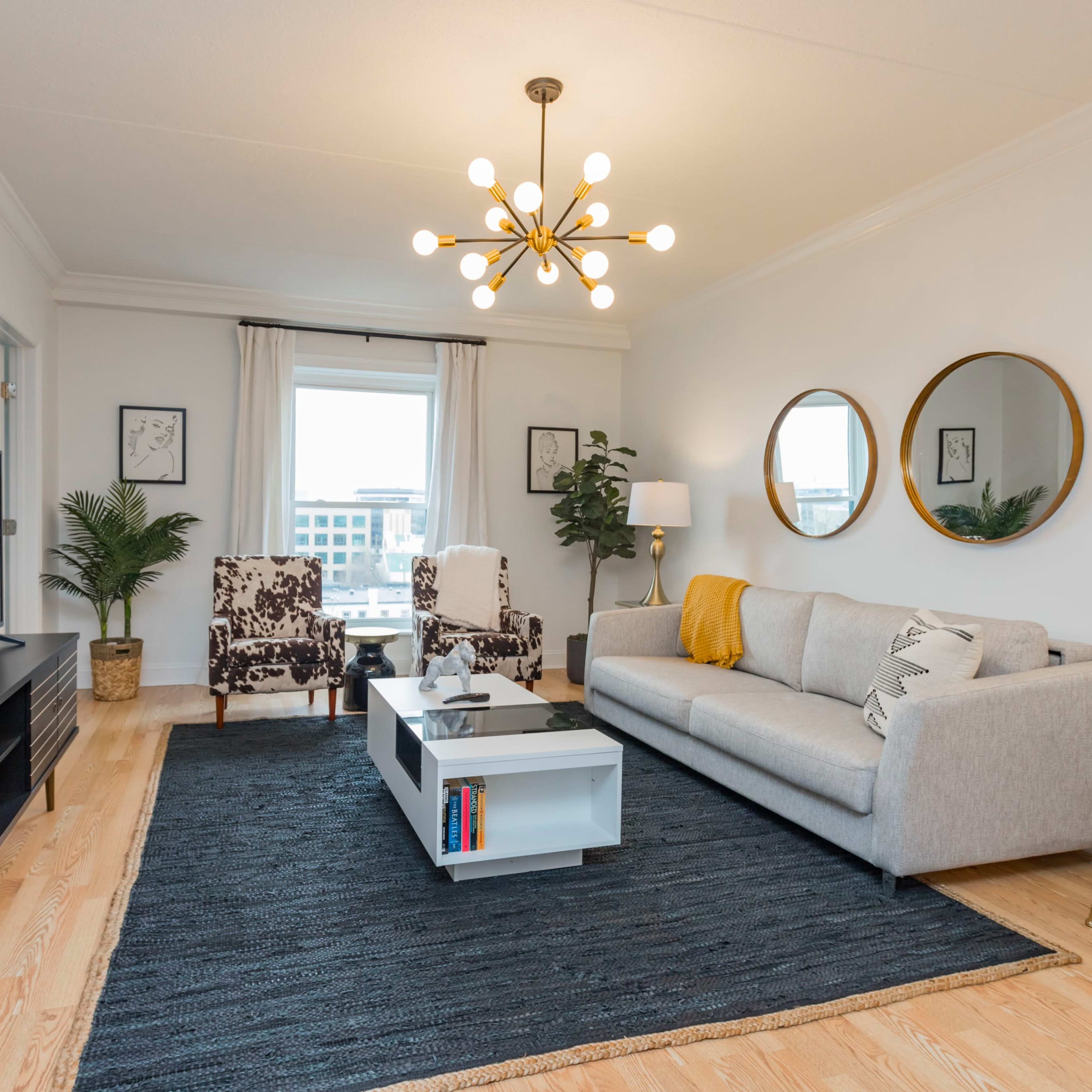 A modern living room features a gray sofa, a black TV stand, round mirrors, and patterned chairs, with a light fixture overhead and a large window illuminating the space.