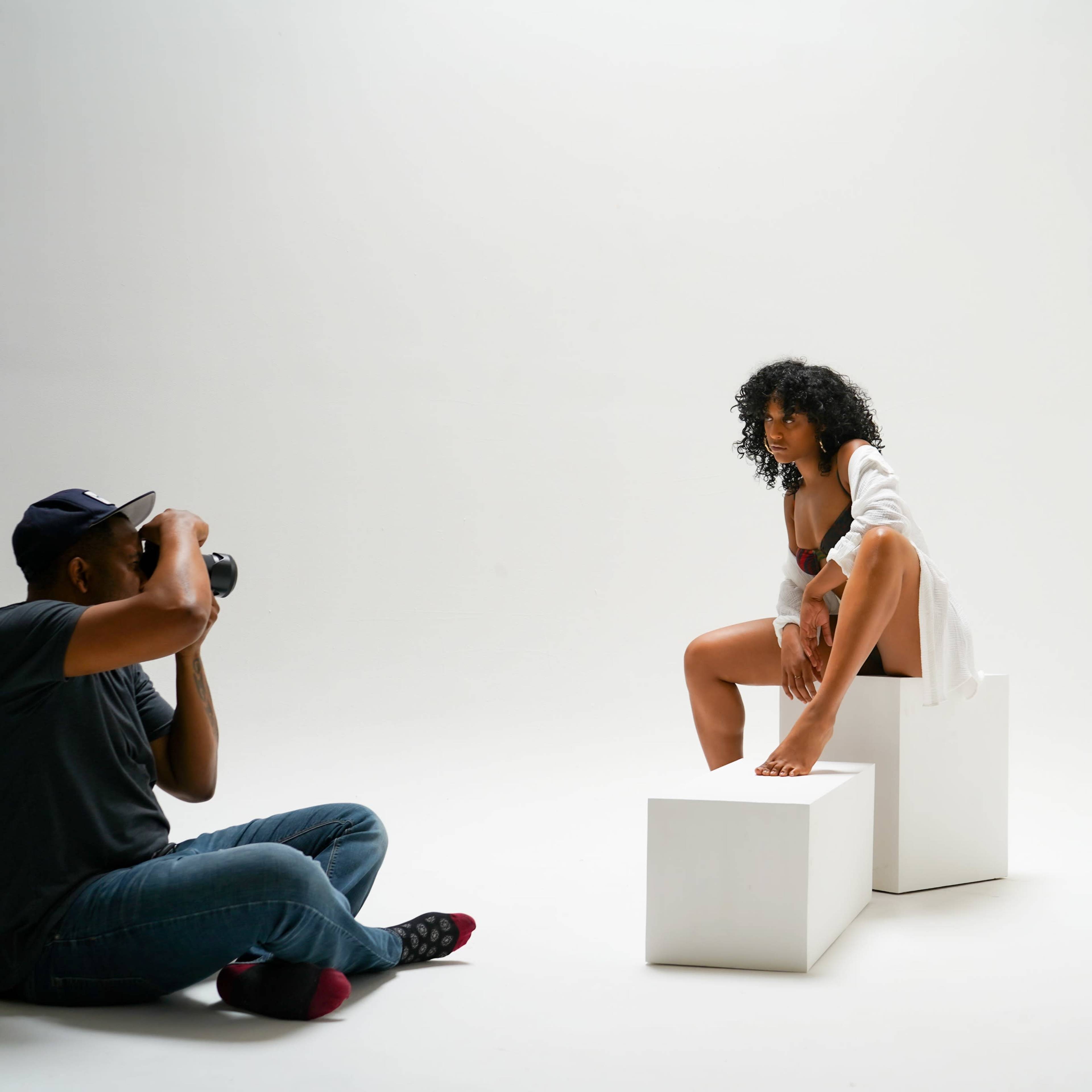 A photographer captures a model posing on a white block in a minimalist studio setting.