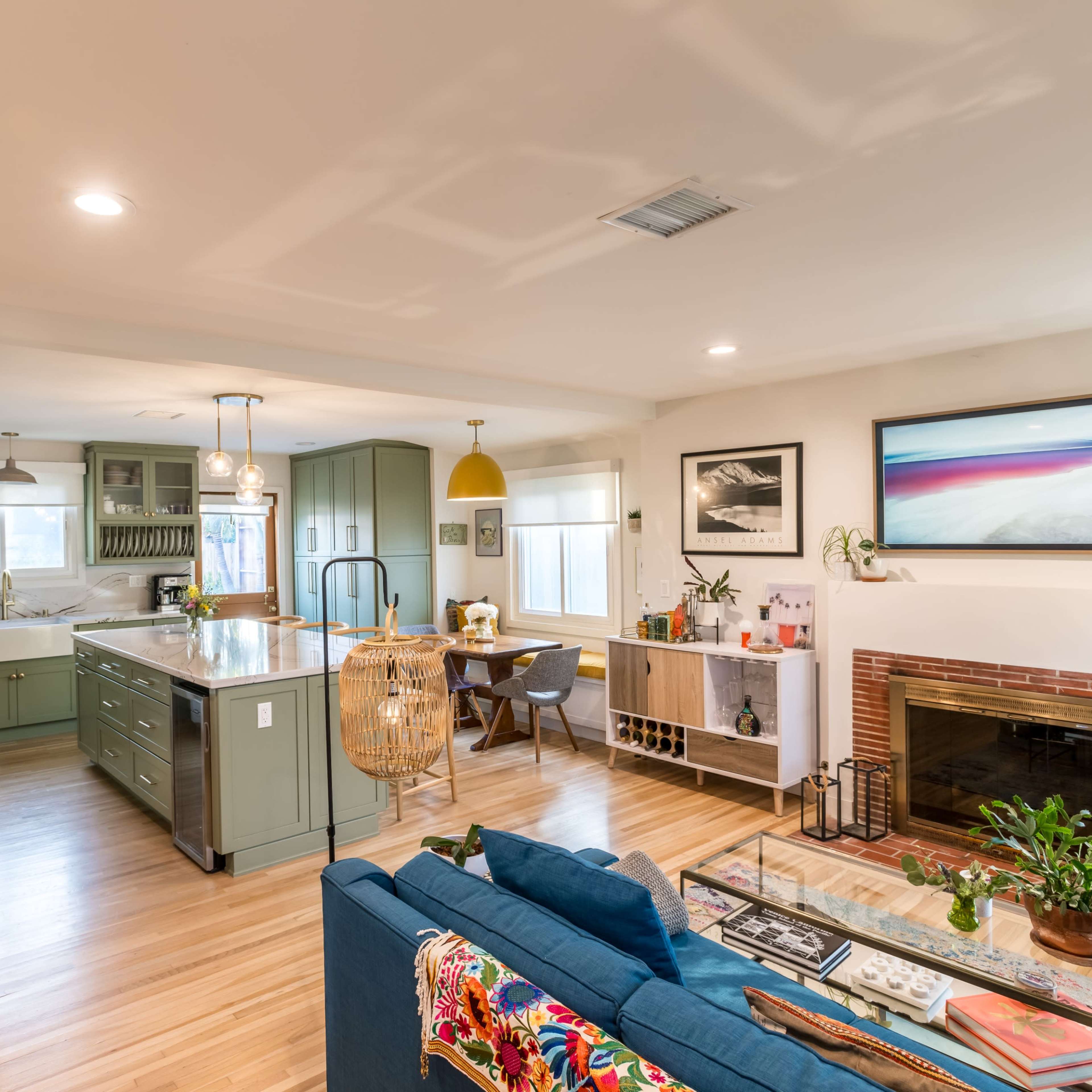 A modern living space featuring a blue sofa, a bookshelf filled with books, a kitchen with green cabinetry, and a fireplace adorned with plants.